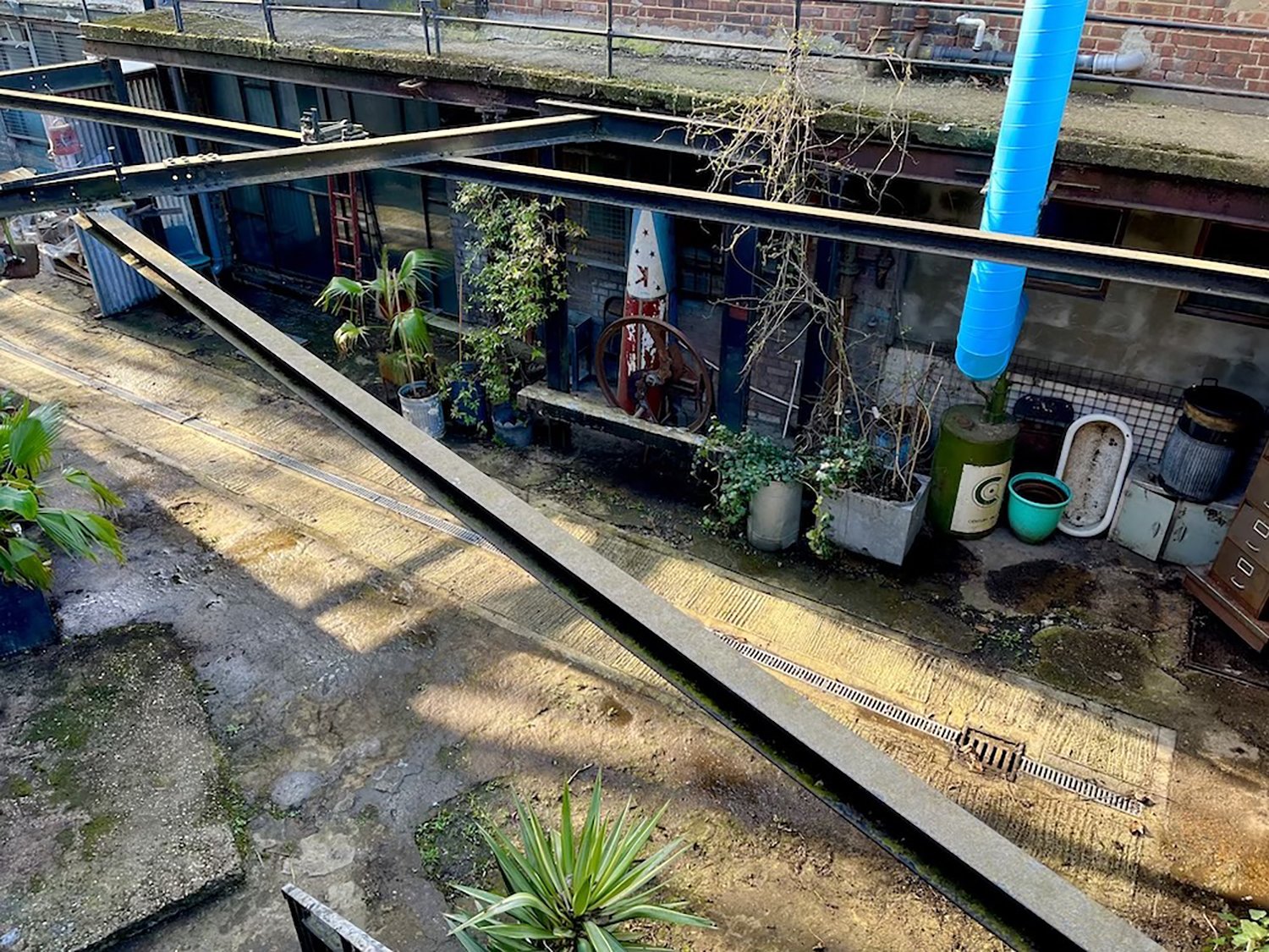 View of an outdoor area with potted plants, a blue vent pipe, a rusted vintage barrel, and various containers and debris."}