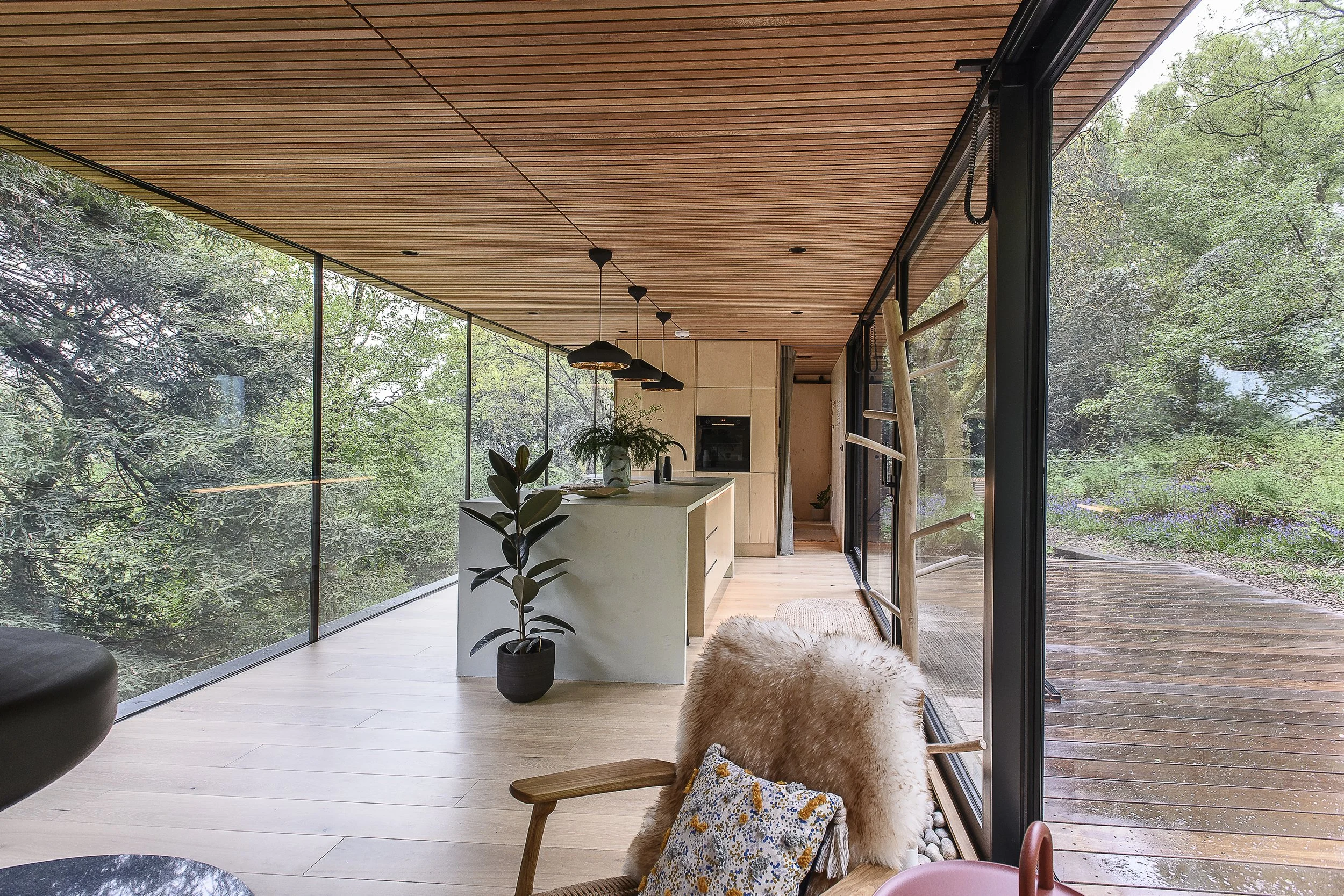 Modern interior with large glass windows overlooking greenery, wooden ceiling, and minimalistic furniture including a cozy chair with a fur throw and pillow, a potted plant, and a kitchen island.