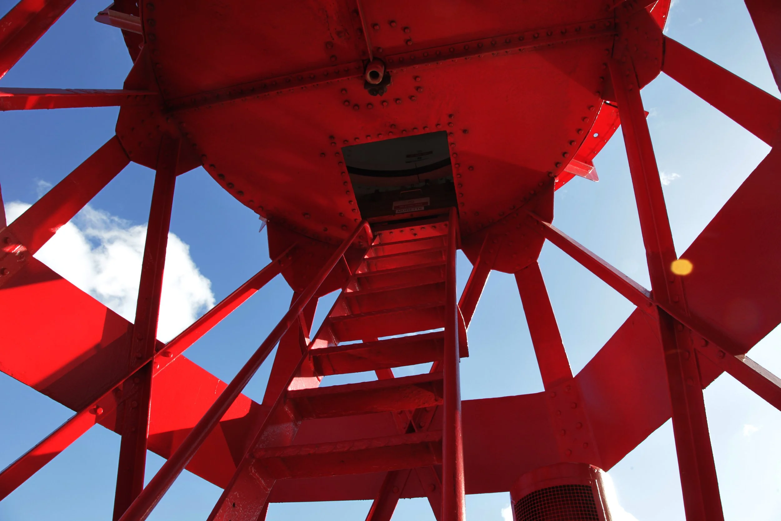 Looking up at a red lighthouse tower with a metal staircase inside, against a blue sky with clouds.