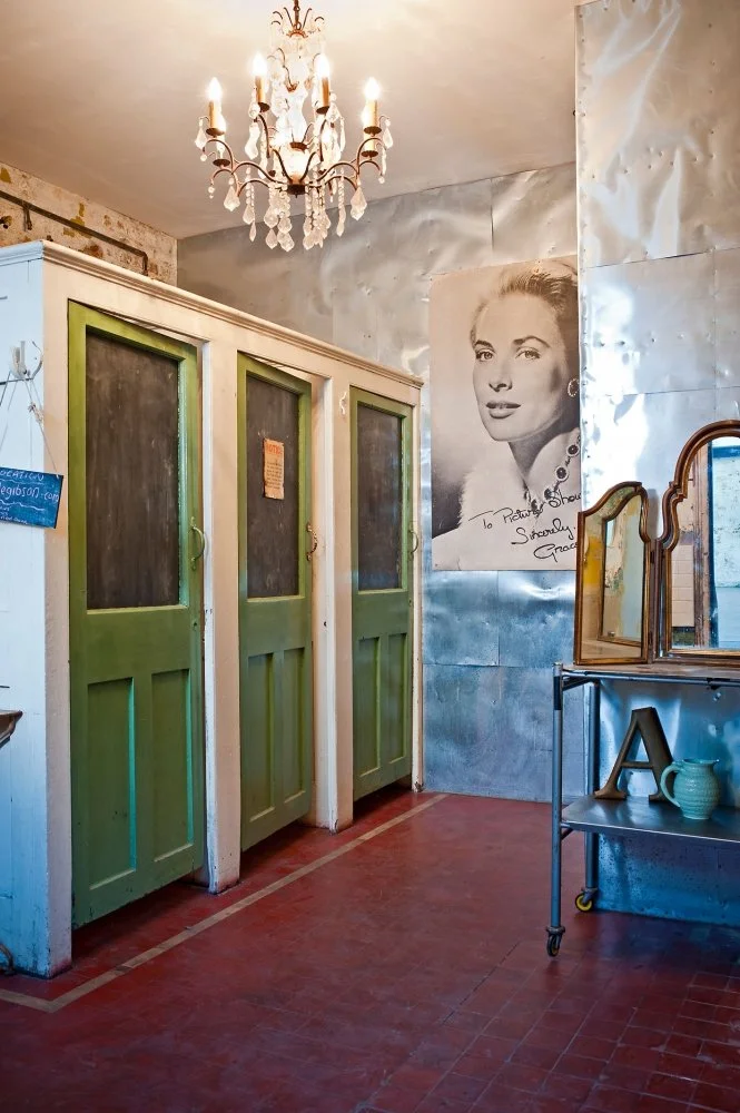 Interior of a vintage-style room with three green-paneled wooden doors, an elegant chandelier hanging from the ceiling, a large black-and-white portrait of a woman on the wall, and a metal table with a mirror, letter A, and a green pitcher.