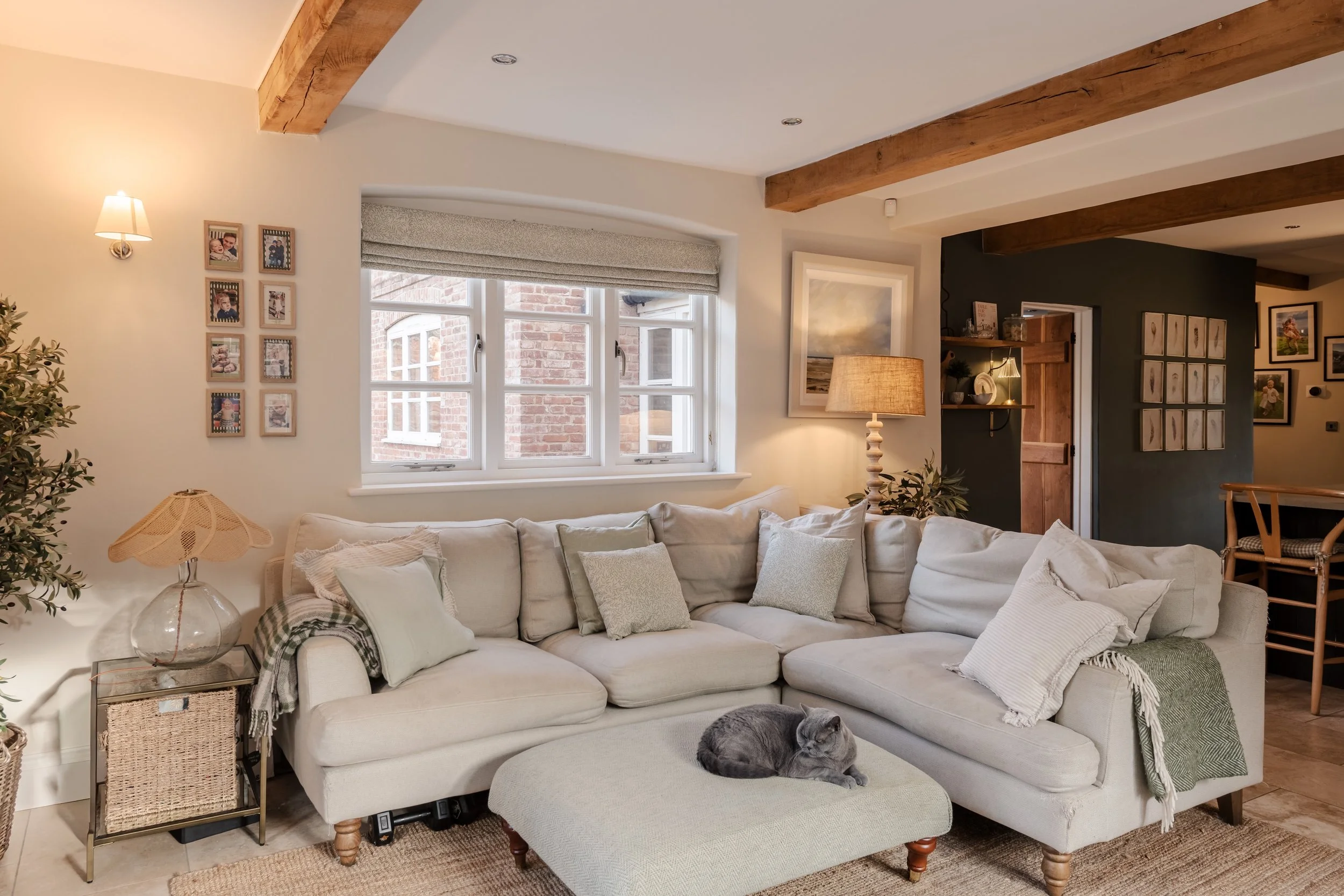Living room with a large white sectional sofa, a grey cat lying on an ottoman, and decor including pillows, lamps, and wall art.