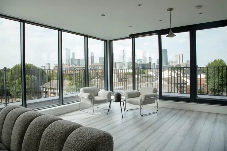 Modern living room with large floor-to-ceiling windows overlooking a city skyline, furnished with white armchairs, a small black table, and a gray couch.