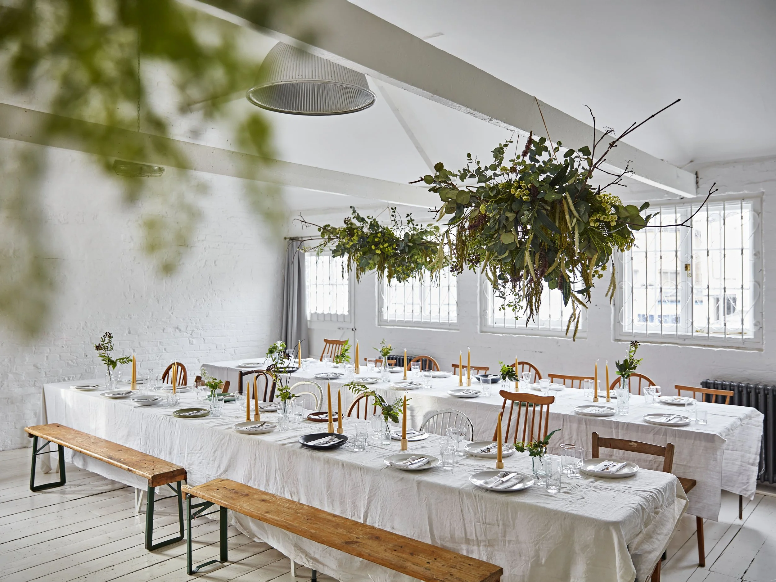 A long table set for a meal with white tablecloths, plates, silverware, glasses, small plant centerpieces, and tall yellow candles. The room has white brick walls, large windows with bars, and hanging greenery overhead.