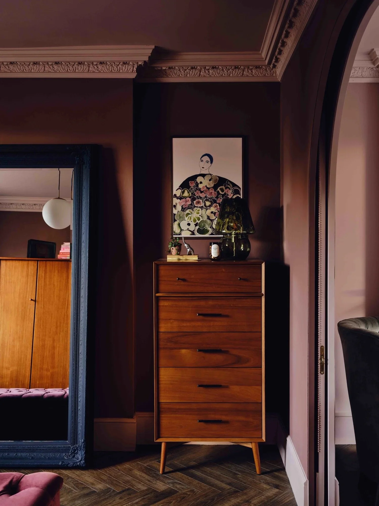 A wooden dresser with six drawers and black handles in a dimly lit room. On top of the dresser are decorative items including a black lamp, a small plant, a candle, and a botanical artwork of a woman with flowers in her hair.