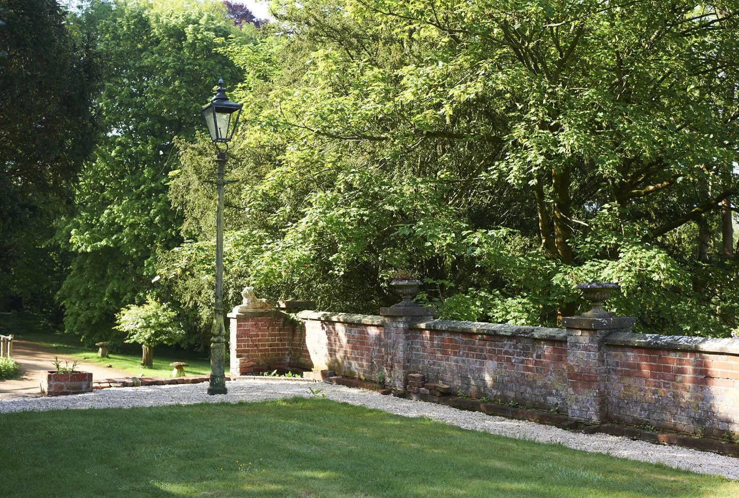 A countryside scene featuring a brick wall with decorative pillars, a vintage black street lamp, lush green trees, well-maintained grass, and a gravel path.