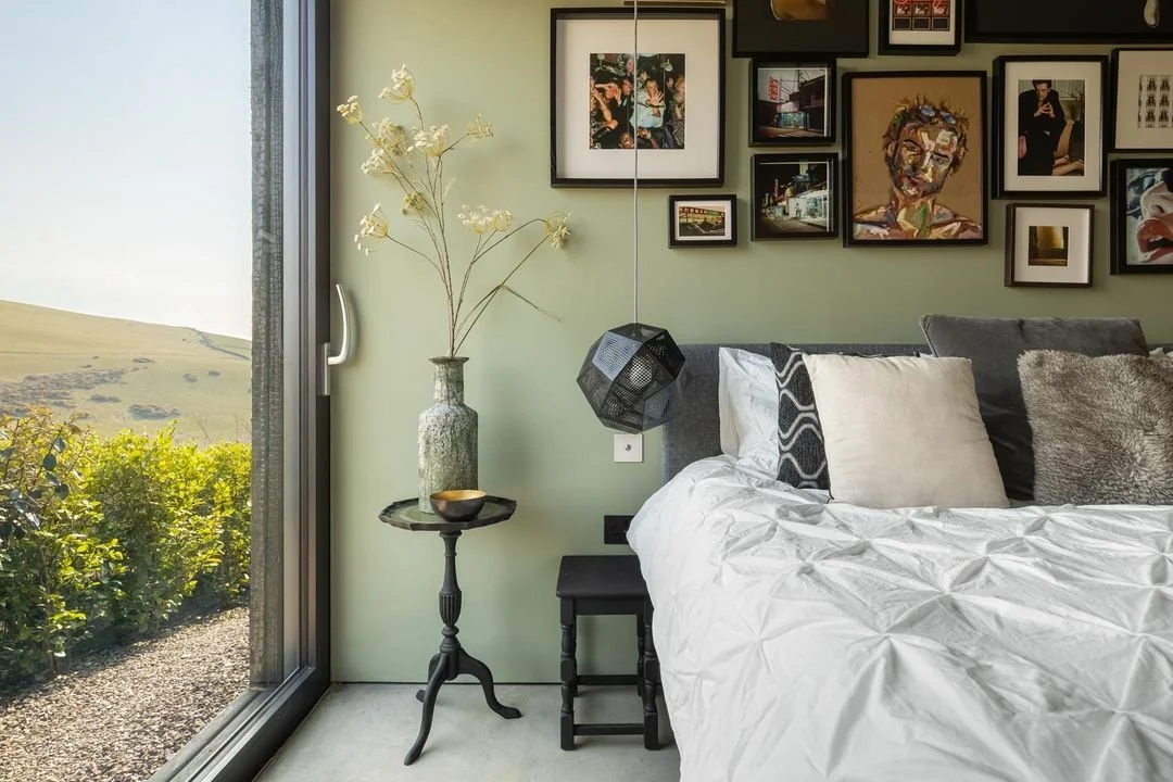 Interior of a bedroom with a bed, pillows, and a gallery wall of framed artwork. Beside the bed is a black side table with a tall vase containing flowers, and a black geometric hanging light. A large window shows a scenic landscape outside.