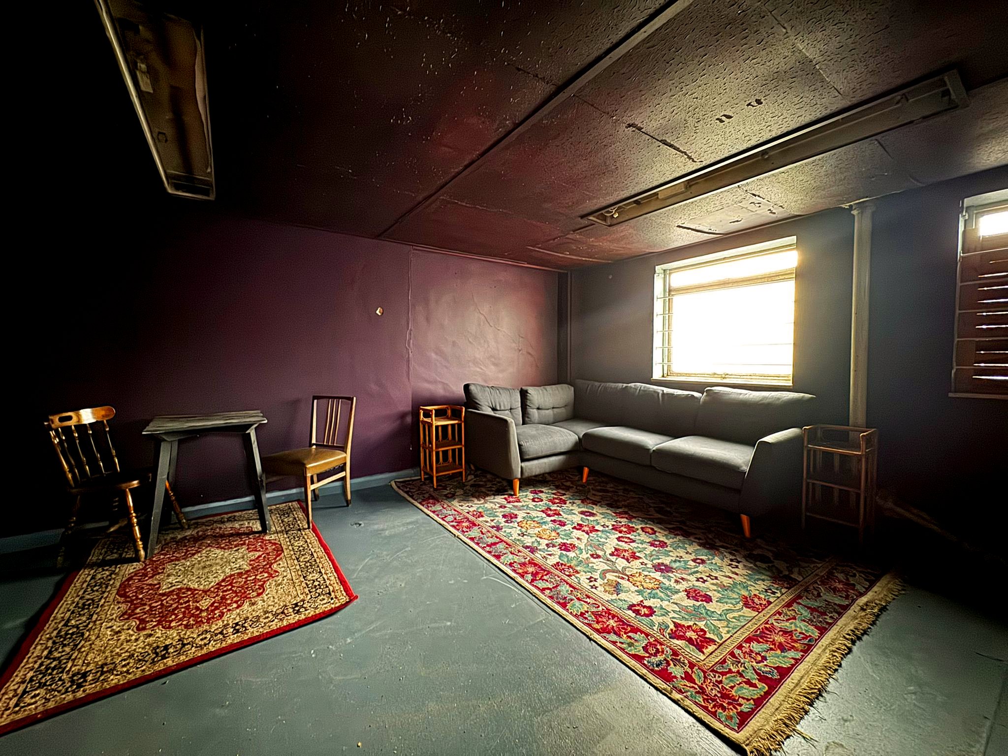 A room with purple walls, a gray sectional sofa, a wooden chair, and several oriental-style rugs on a gray concrete floor. There is a window with blinds on the bright wall, and a ceiling vent.