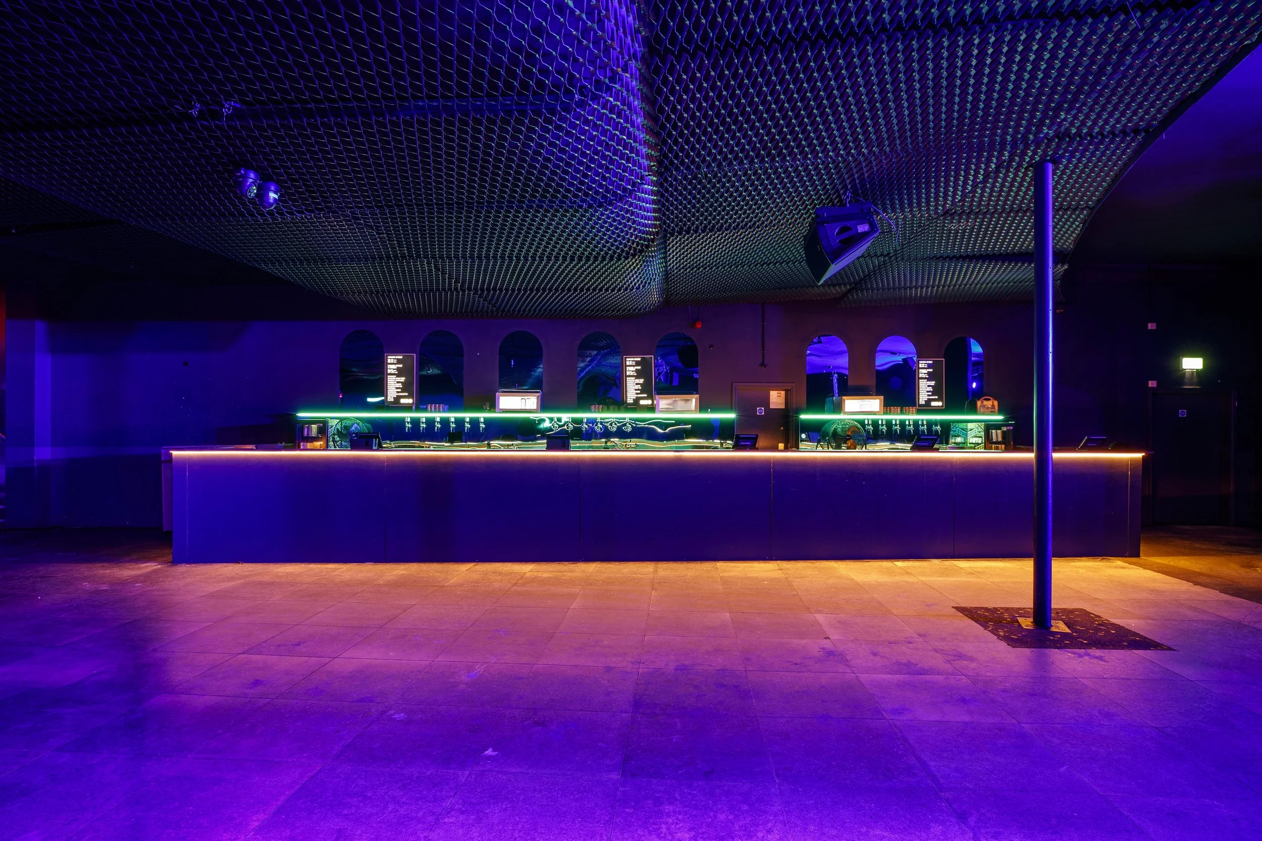 Empty nightclub bar with neon lighting, dark ceiling with a grid pattern, and a single pole in the foreground.