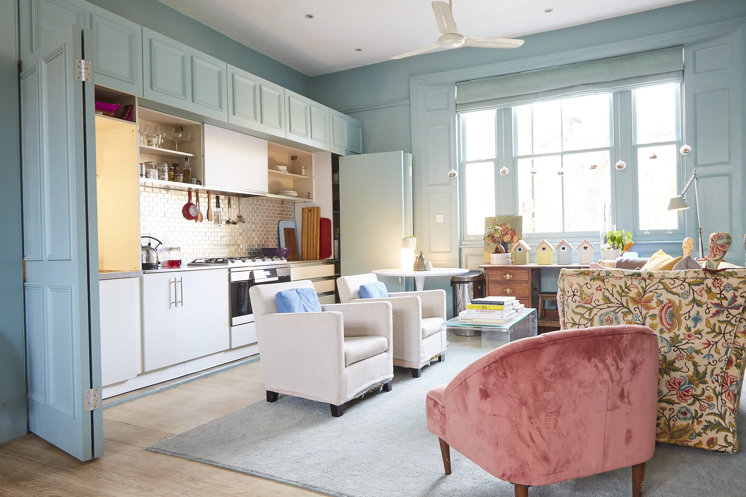 A bright, open-plan living room and kitchen area with light blue walls and cabinetry, white armchairs with blue cushions, a pink velvet armchair, a floral-patterned sofa, and a window with sunlight streaming in. The kitchen features a stovetop, oven,