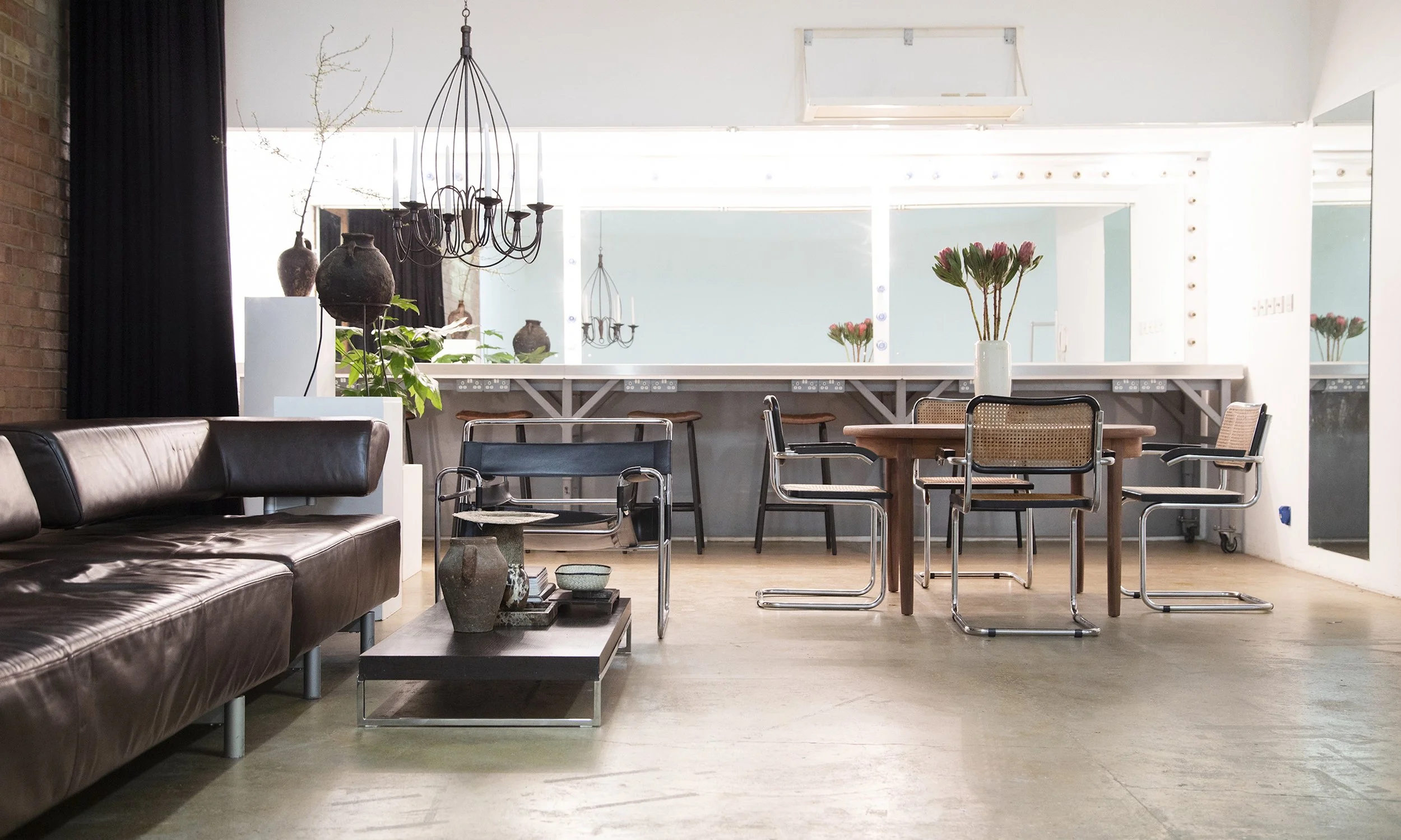 Interior of a modern living space with a dark leather sofa, various chairs around a wooden table, and decorative vases and plants. Large windows and chandeliers provide natural and ambient lighting.