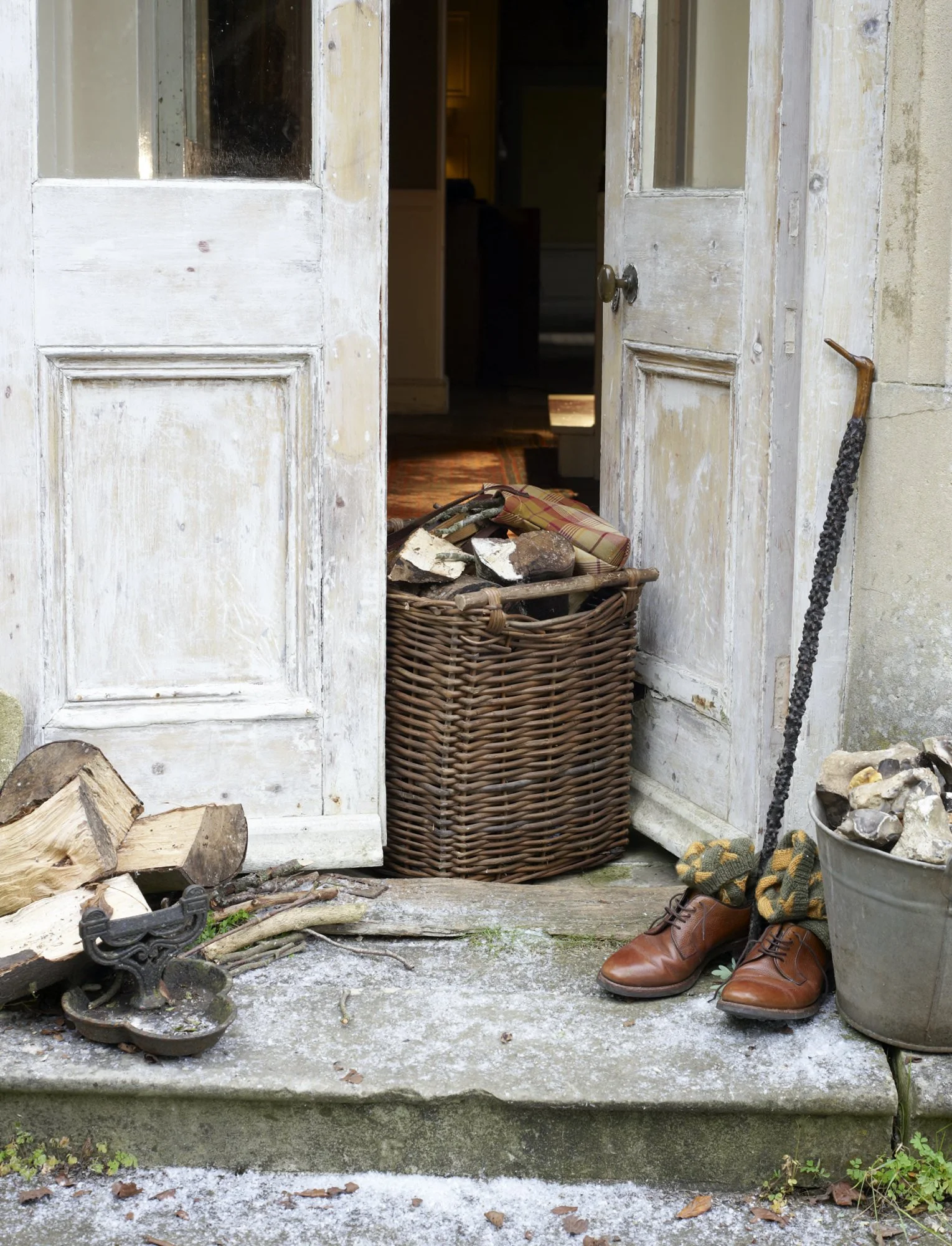 Open front door revealing a pile of firewood and a basket filled with kindling, with shoes and gardening tools outside.