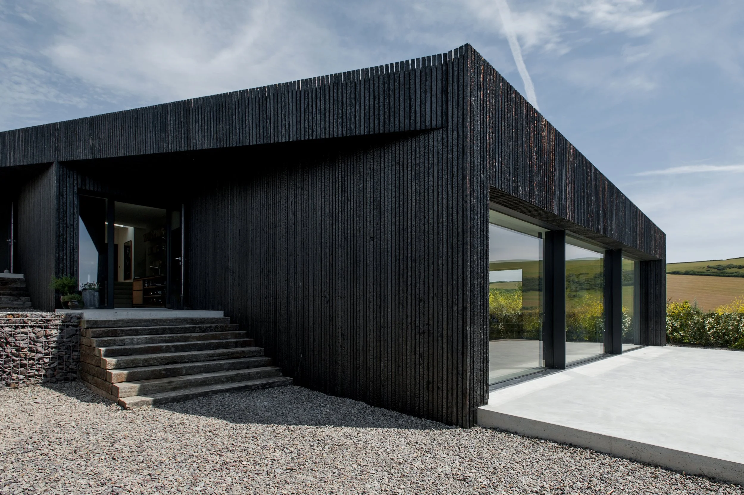 Modern black house with steps leading to entrance, large glass windows, and a gravel yard, set against a partly cloudy sky.
