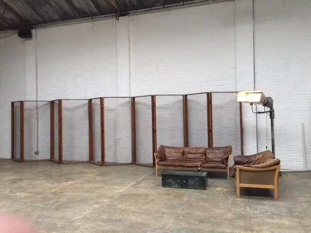 Empty indoor industrial space with a temporary fencing, vintage couch, armchair, side table, and a large studio light.