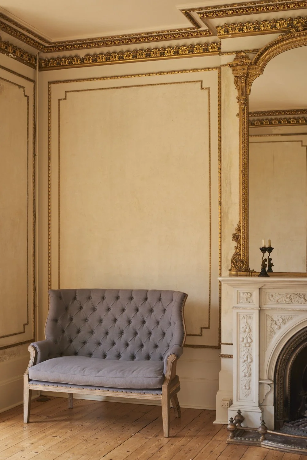 An elegant room corner with a tufted gray sofa, ornate cream and gold wall moldings, a white fireplace with detailed carvings, and a small black candle holder with three white candles on the mantel. The floor is wooden.