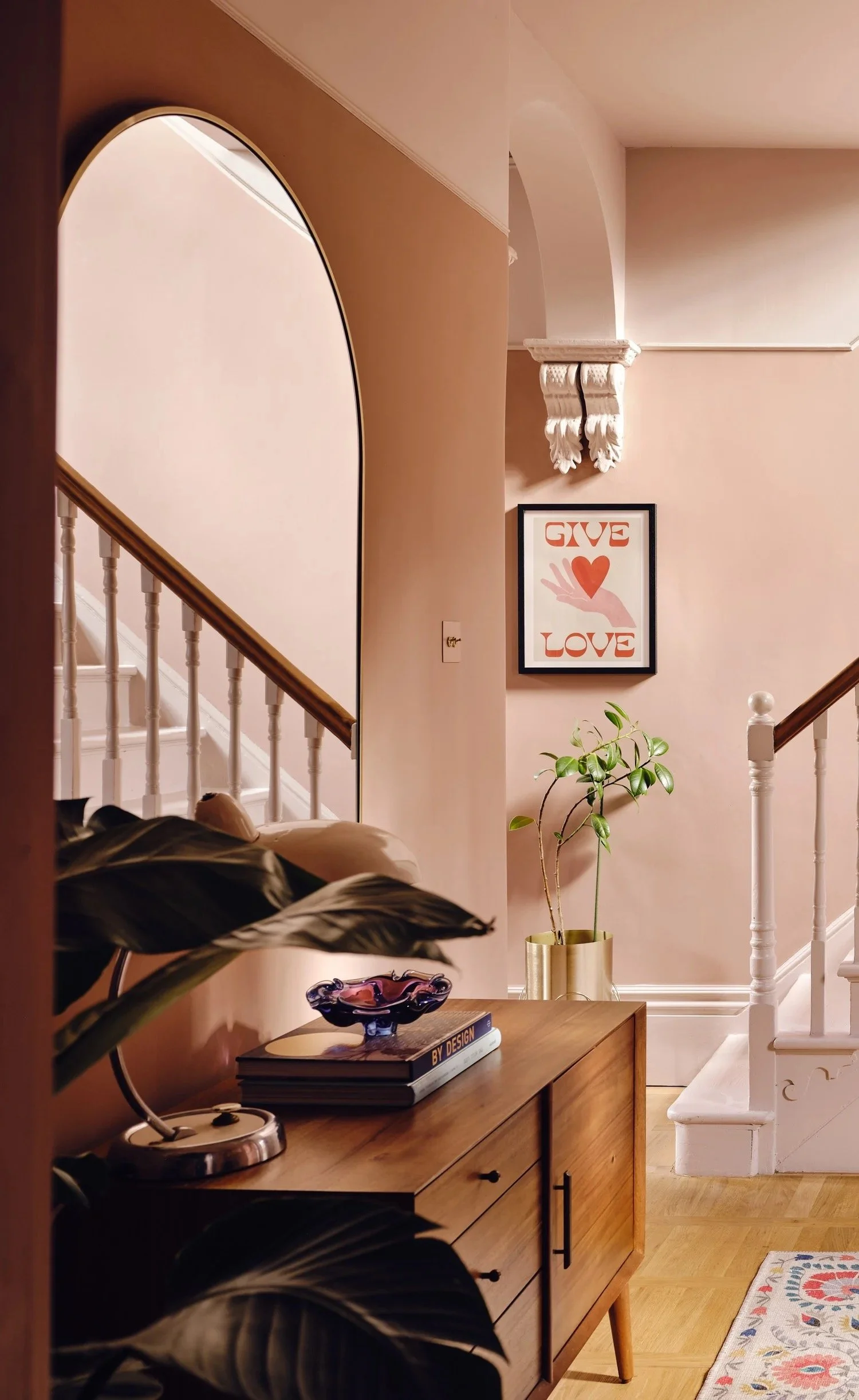Interior view of a cozy home hallway with a wooden dresser, decorative glass bowl, a book titled 'BY DESIGN,' potted plant in a gold planter, framed artwork with the words 'GIVE LOVE' and a hand holding a heart, staircase with white banister, and war