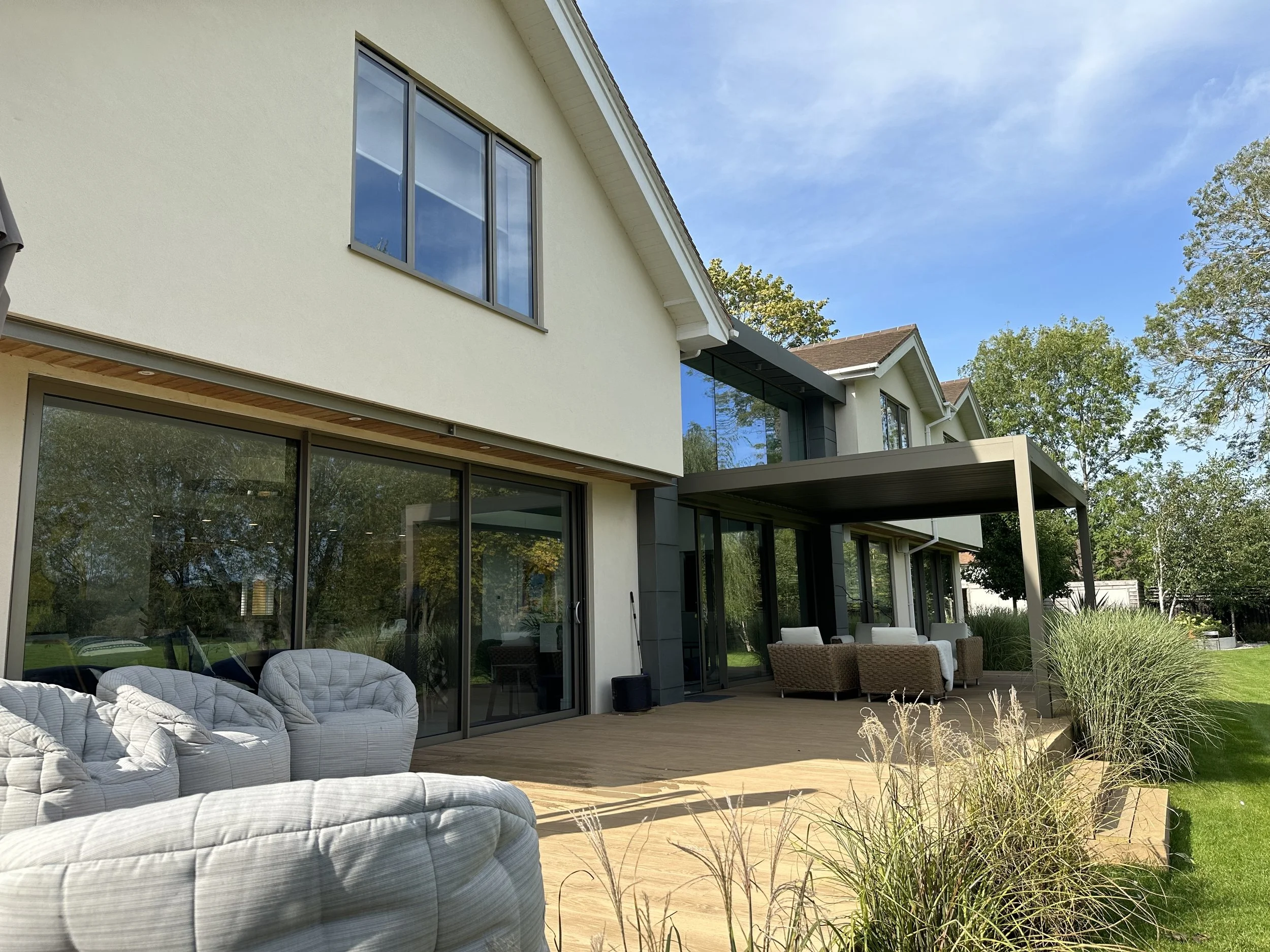 Modern house with large sliding glass doors, outdoor seating on wooden deck, and lush green yard with trees under a blue sky.