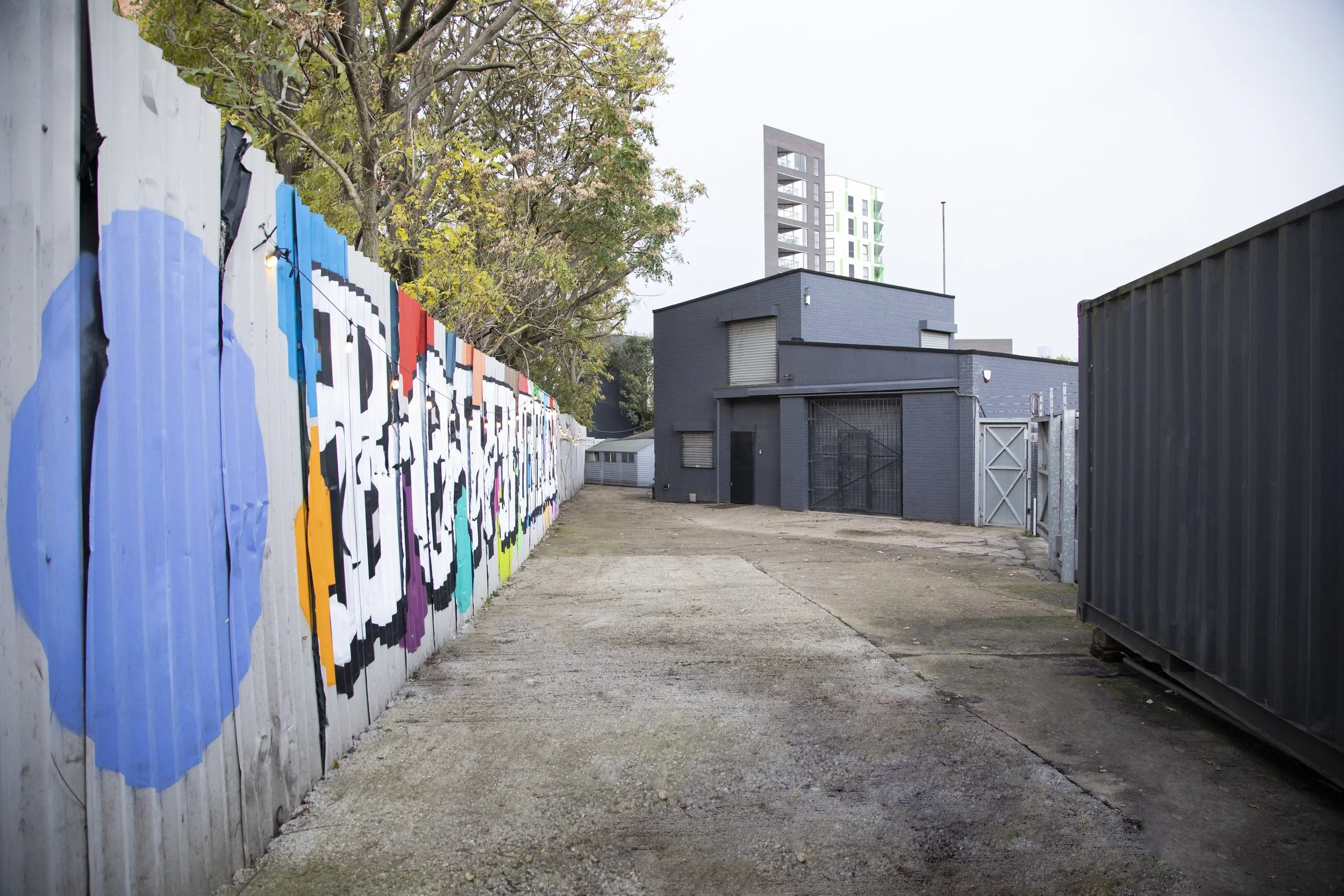 An alleyway with a concrete path, a graffiti-painted fence on the left, a dark gray building with a metal gate at the end, a black shipping container on the right, trees overhead, and a tall modern building in the background.