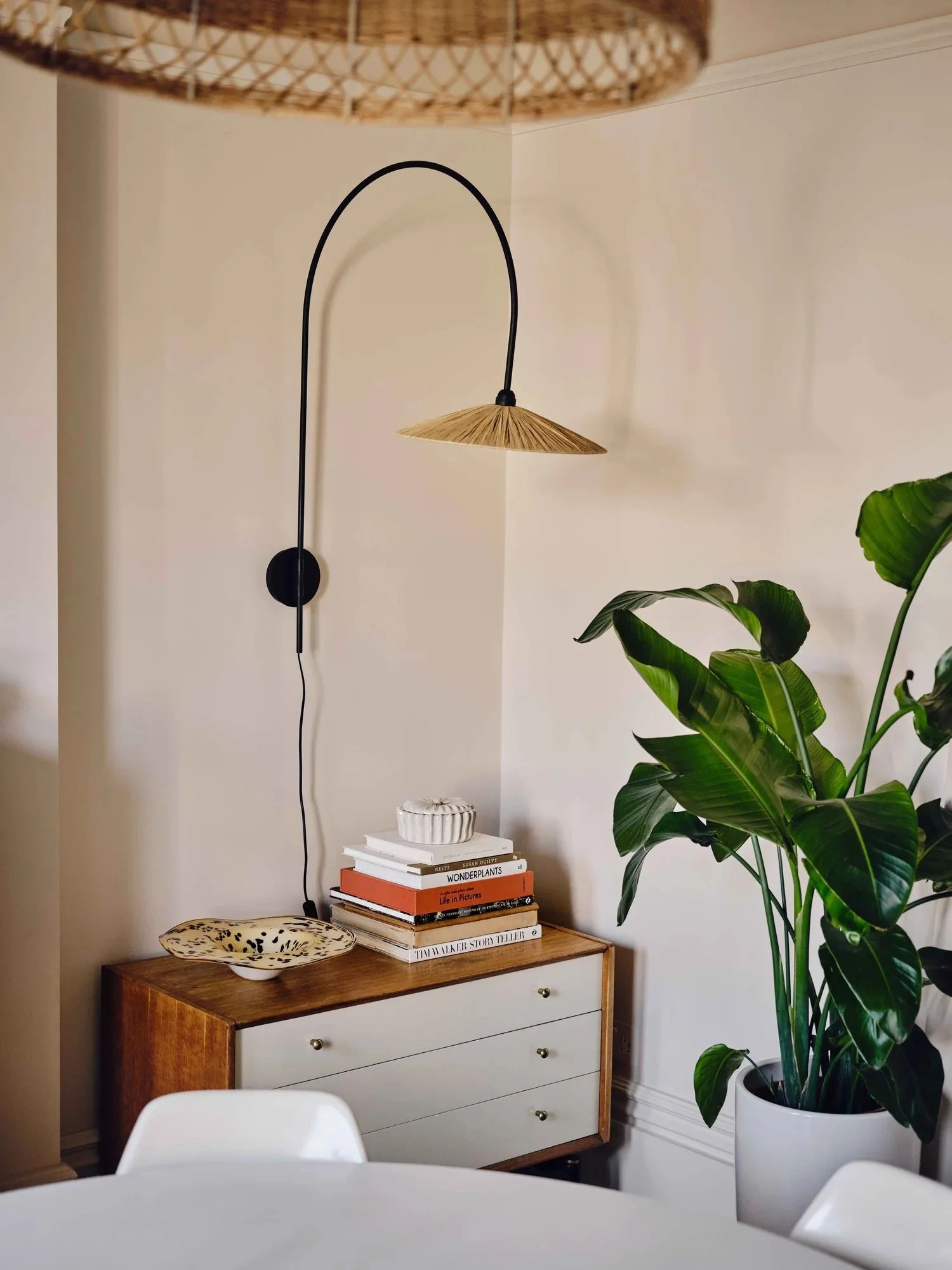Interior of a room with a wooden sideboard against a white wall. A large plant in a white pot is on the right. Above the sideboard is a black wall-mounted lamp with a woven shade. On the sideboard, there is a stack of books and a decorative dish.