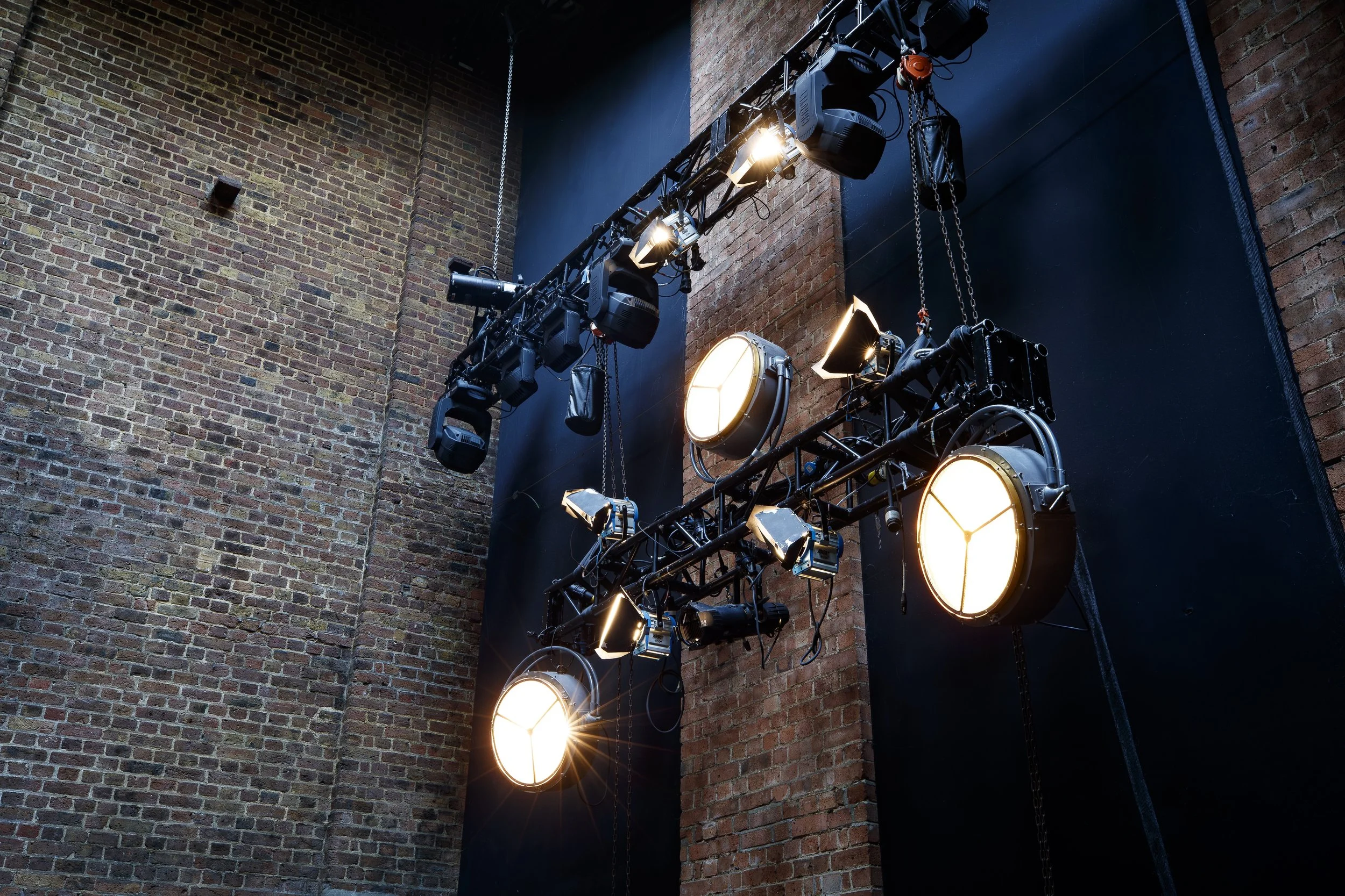 Stage lighting equipment hanging from a black metal truss against a brick wall.