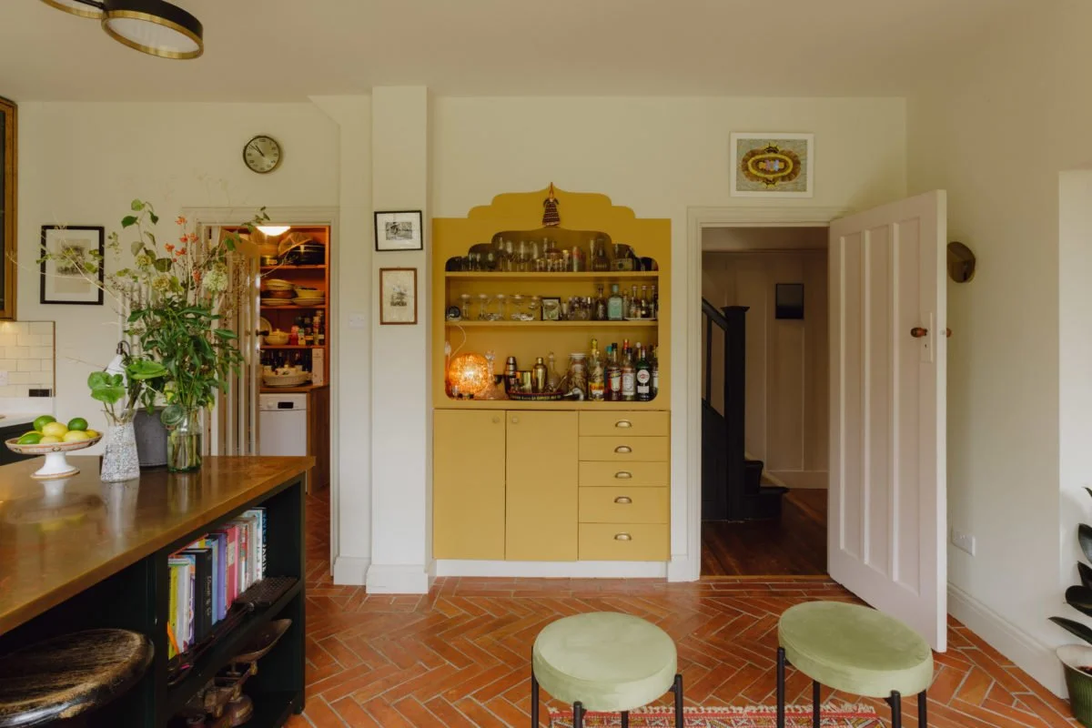 Living room with a yellow bar cabinet, opened white door, stairs, and a bookshelf with books and plants.