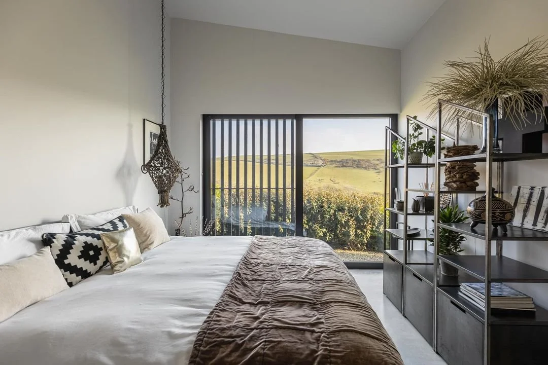 Modern bedroom with a large bed, decorative pillows, a shelf with plants and decorative items, and large sliding glass doors leading to a scenic outdoor view of rolling hills and greenery.