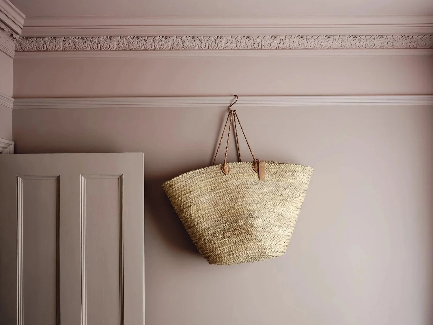 A woven straw basket hanging upside down from a hook on the pink ceiling of a room, next to a slightly open white door with decorative molding.