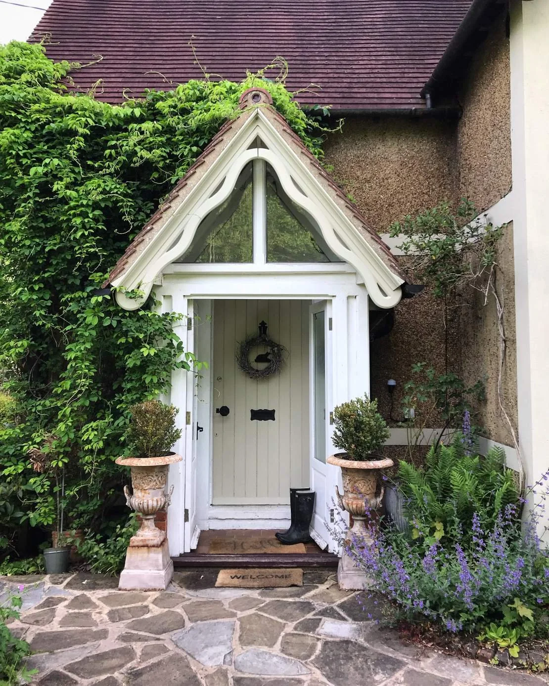 Front entrance of a small house with a decorative white door and a gabled porch, surrounded by lush green plants and potted shrubs, with a stone pathway leading to the door.