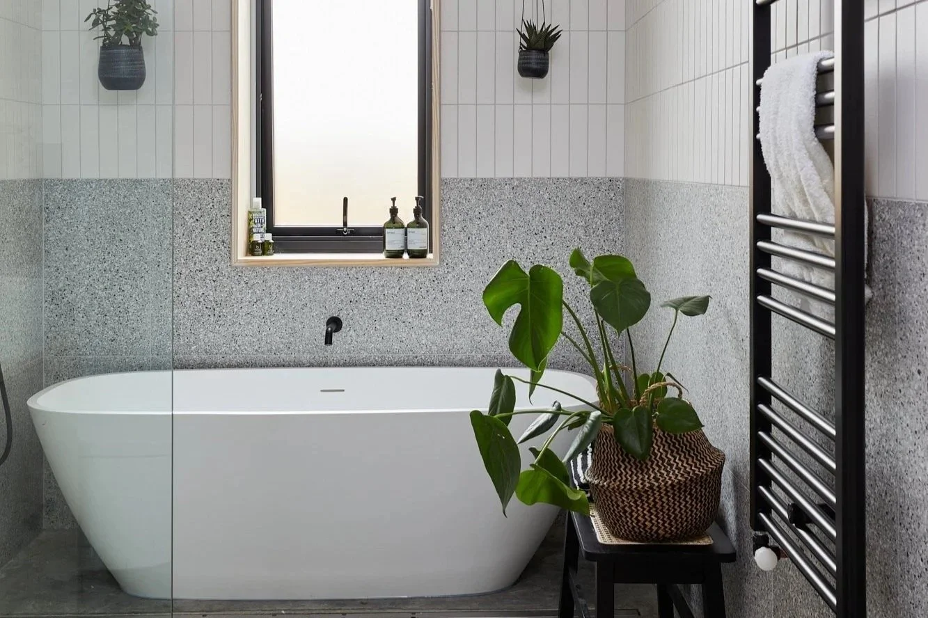 Modern bathroom with a white bathtub, a window with frosted glass, black soap dispensers on the windowsill, a tall black towel rack with a white towel, and a potted green plant on a small black stool.