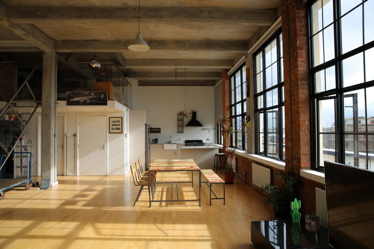 Spacious industrial-style loft apartment with large windows, exposed brick walls, wooden flooring, modern kitchen, dining table with chairs, and potted plants.