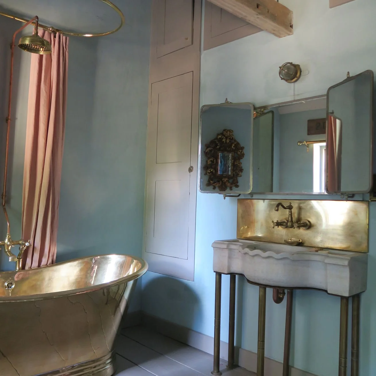 Vintage bathroom with gold bathtub, pink curtain, and an ornate mirror, featuring a brass sink and a framed mirror on light blue walls.