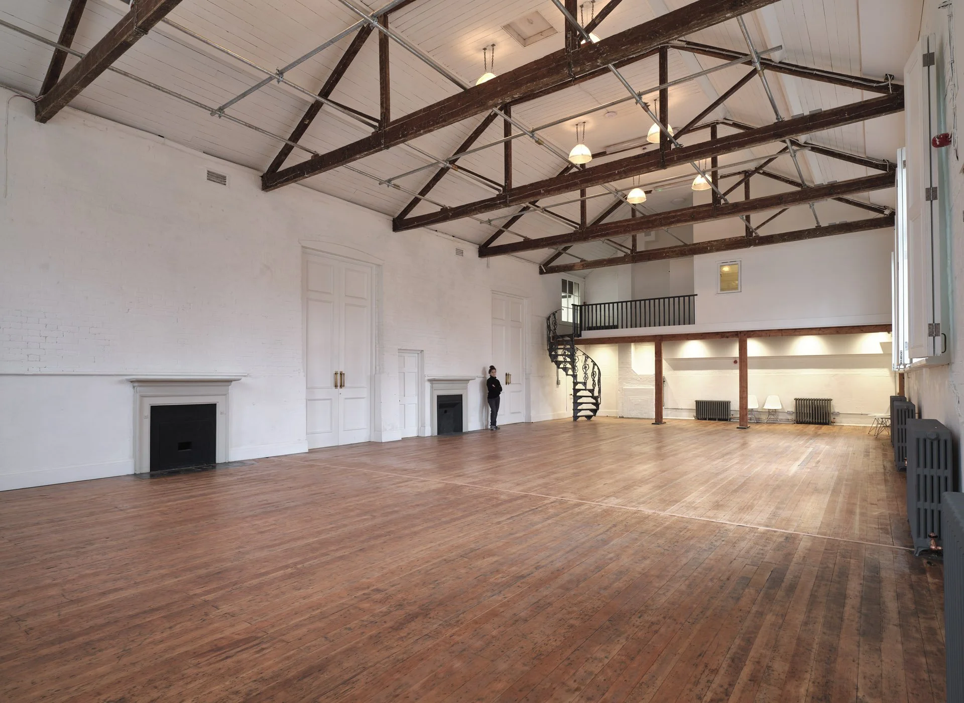 Spacious, empty room with wooden floors, white walls, exposed wooden beams on the ceiling, and a spiral staircase leading to a loft area.