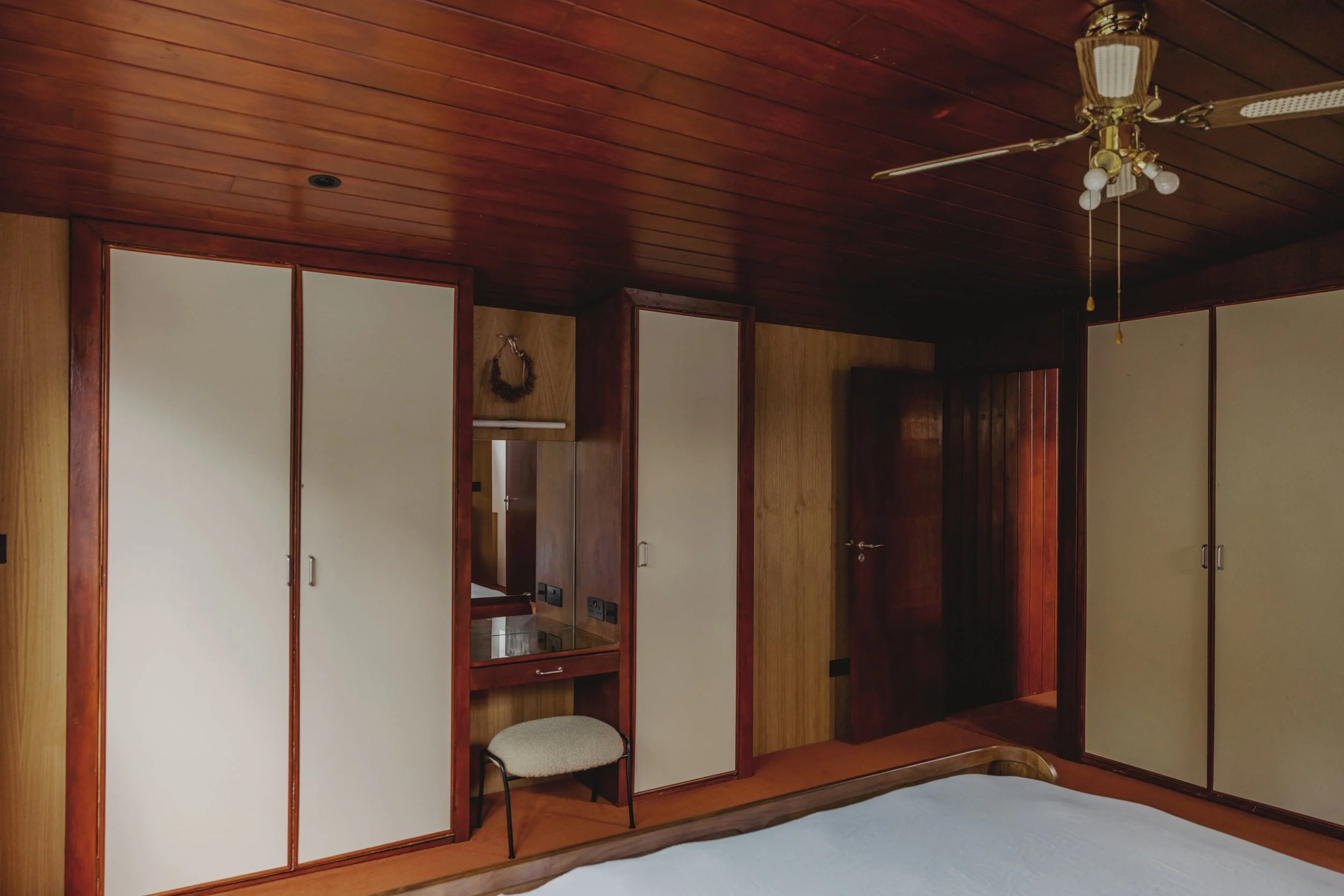 A bedroom with wood-paneled ceiling and walls, fixtures include a ceiling fan with four blades and four light bulbs. The room has built-in wardrobes with white doors, a small dressing table with a mirror and a beige cushioned stool, and an open door 