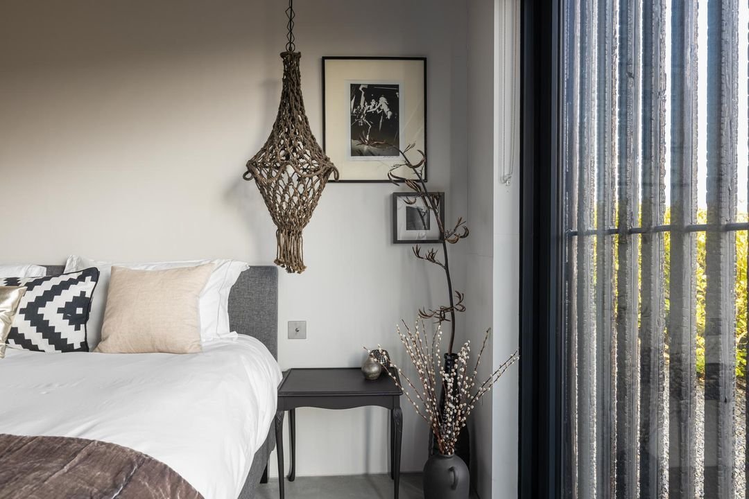 A bedroom corner with a grey bed, decorative pillows, a black nightstand, framed photos on the wall, a macramé wall hanging, a tall vase with branches and pussy willow stems, and large window curtains.
