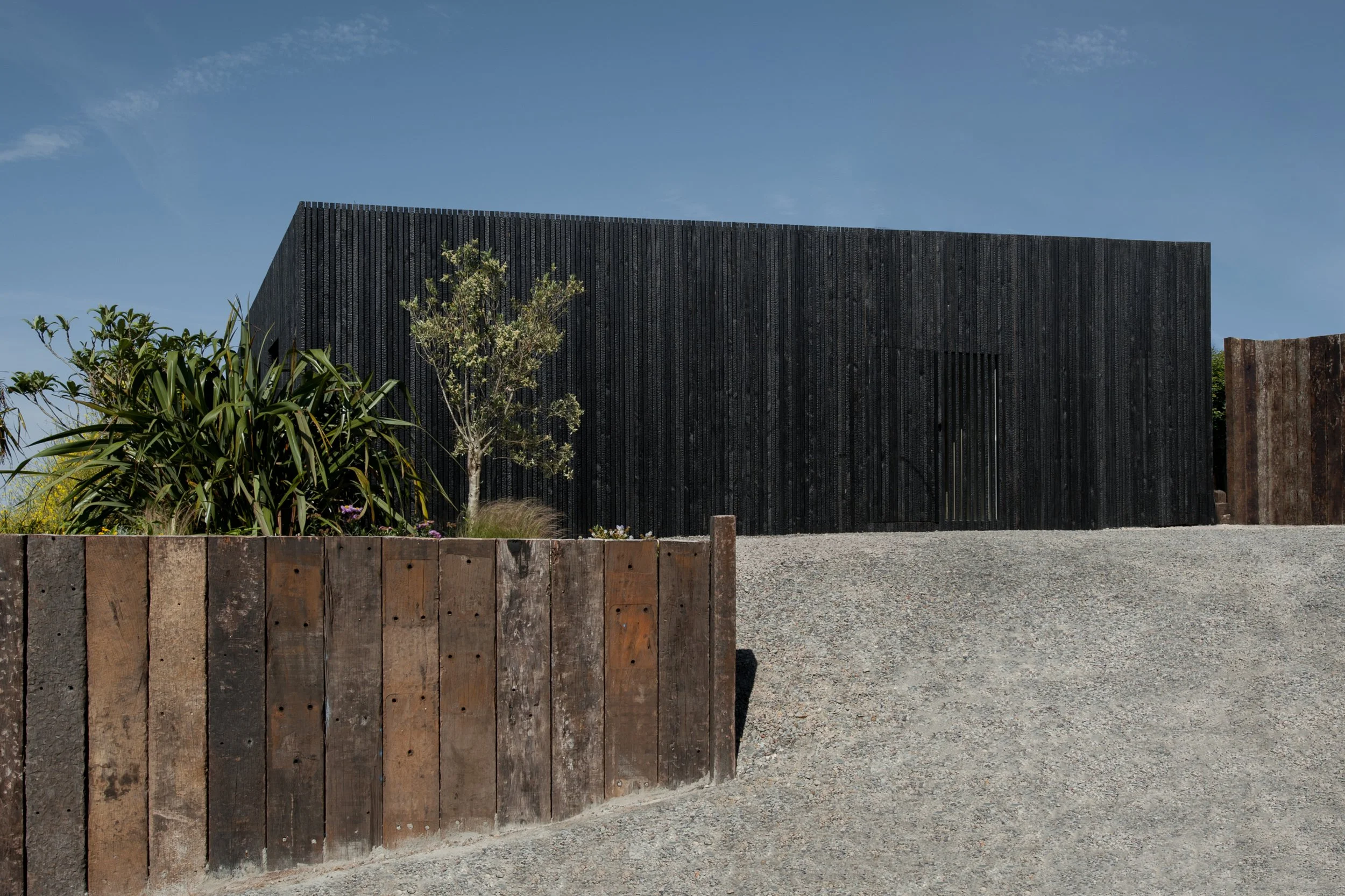 Modern black wooden building on a gravel driveway with a wooden fence and some plants and trees in front, under a blue sky.