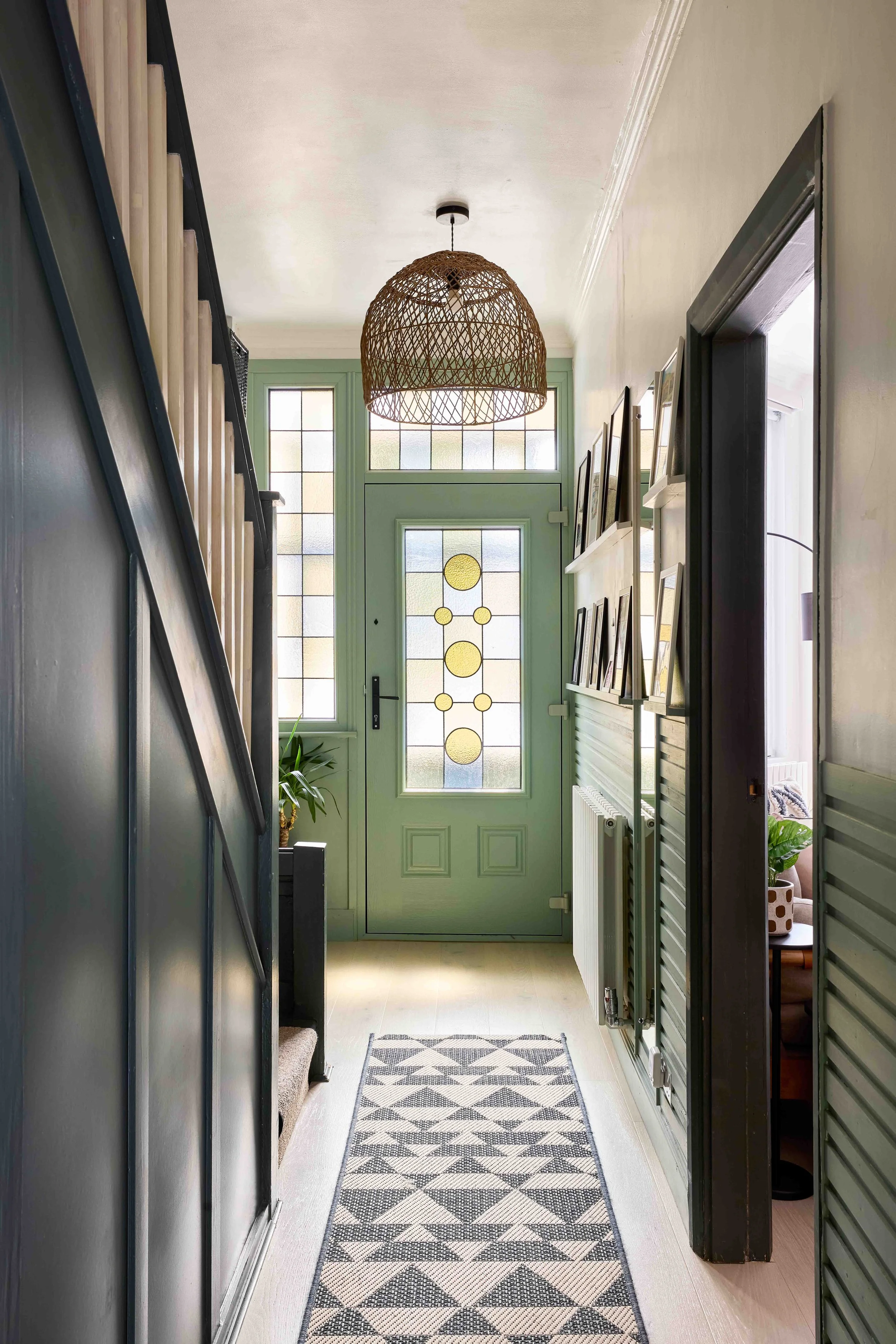 A narrow hallway leading to a door with decorated glass panels. The hallway has a patterned rug on the floor, a black railing on the left, picture frames on the right, and a rattan pendant light hanging from the ceiling.