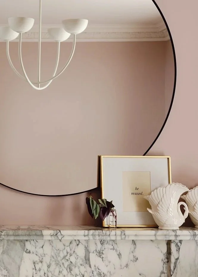 A round mirror hanging above a marble mantle, reflecting a white ceiling chandelier. On the mantle, there is a framed art print with the words 'be mused.' and decorative white seashell sculptures next to a small plant in a glass bottle.