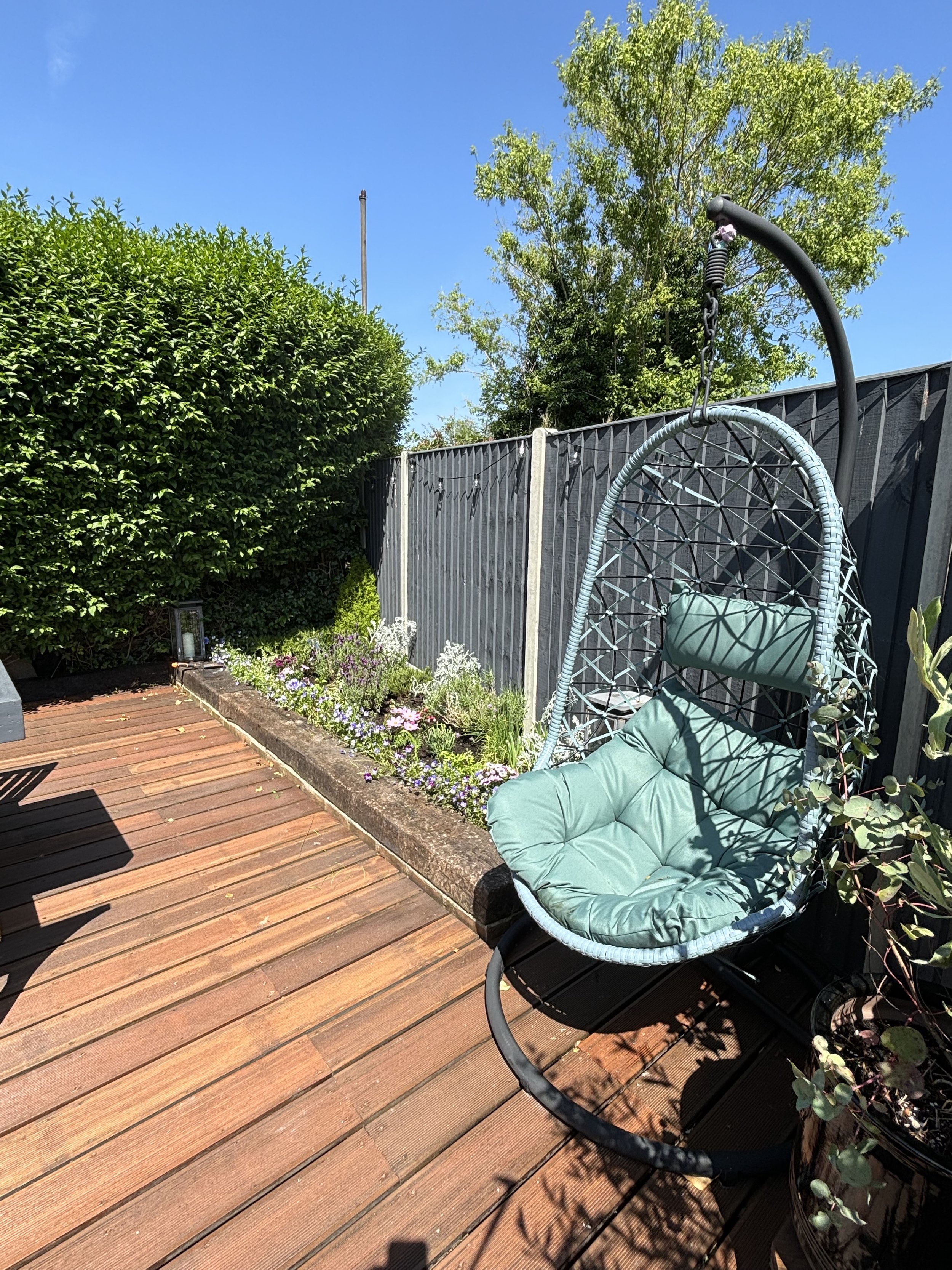 A backyard with a wooden deck, a hanging egg-shaped swing chair with cushions, a flower bed with blooming plants, a tall bush, and a black fence under a clear blue sky.