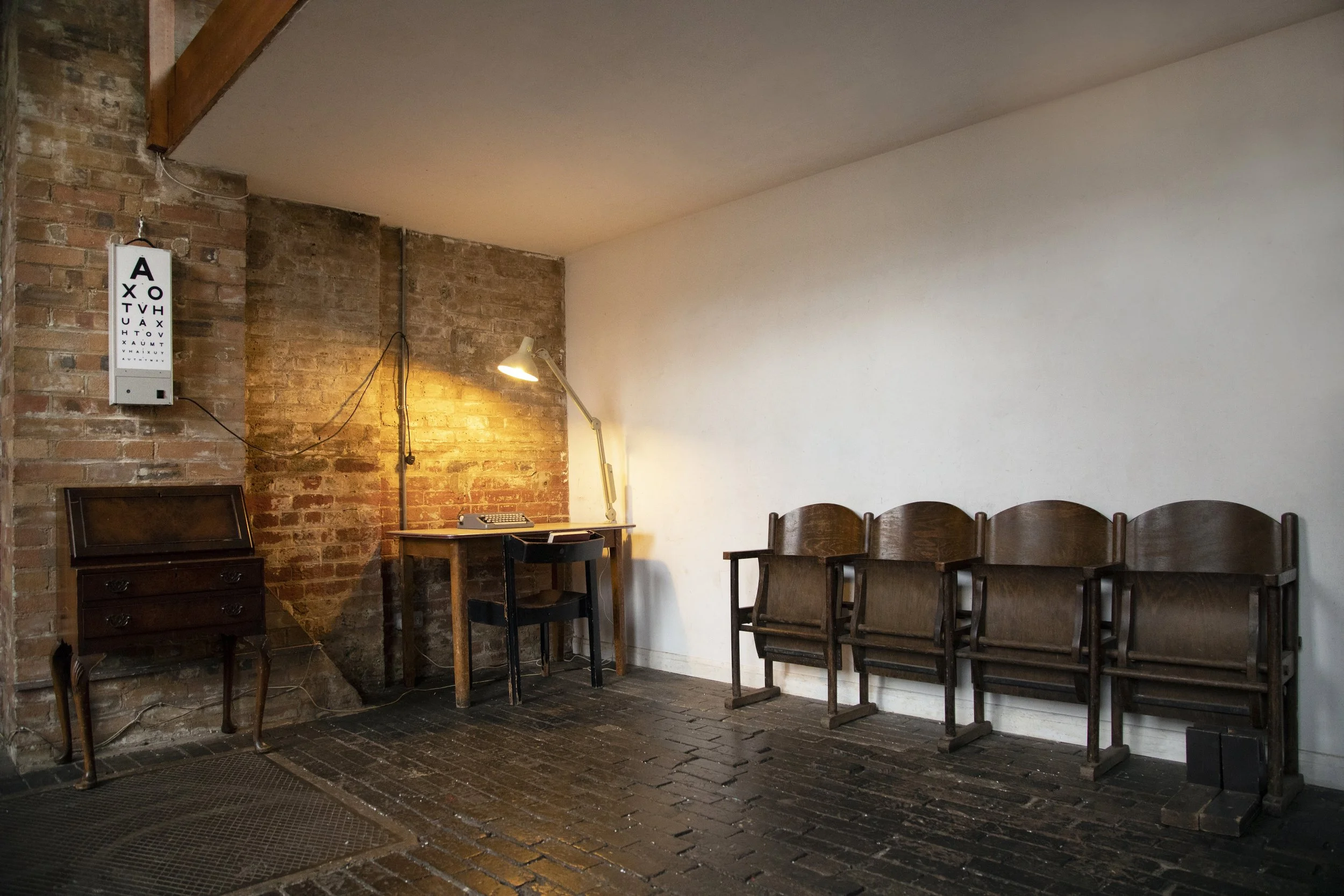 Interior of a minimalist room with a brick wall, wooden furniture, a row of empty chairs, a desk with a lamp, and an eye chart on the wall.