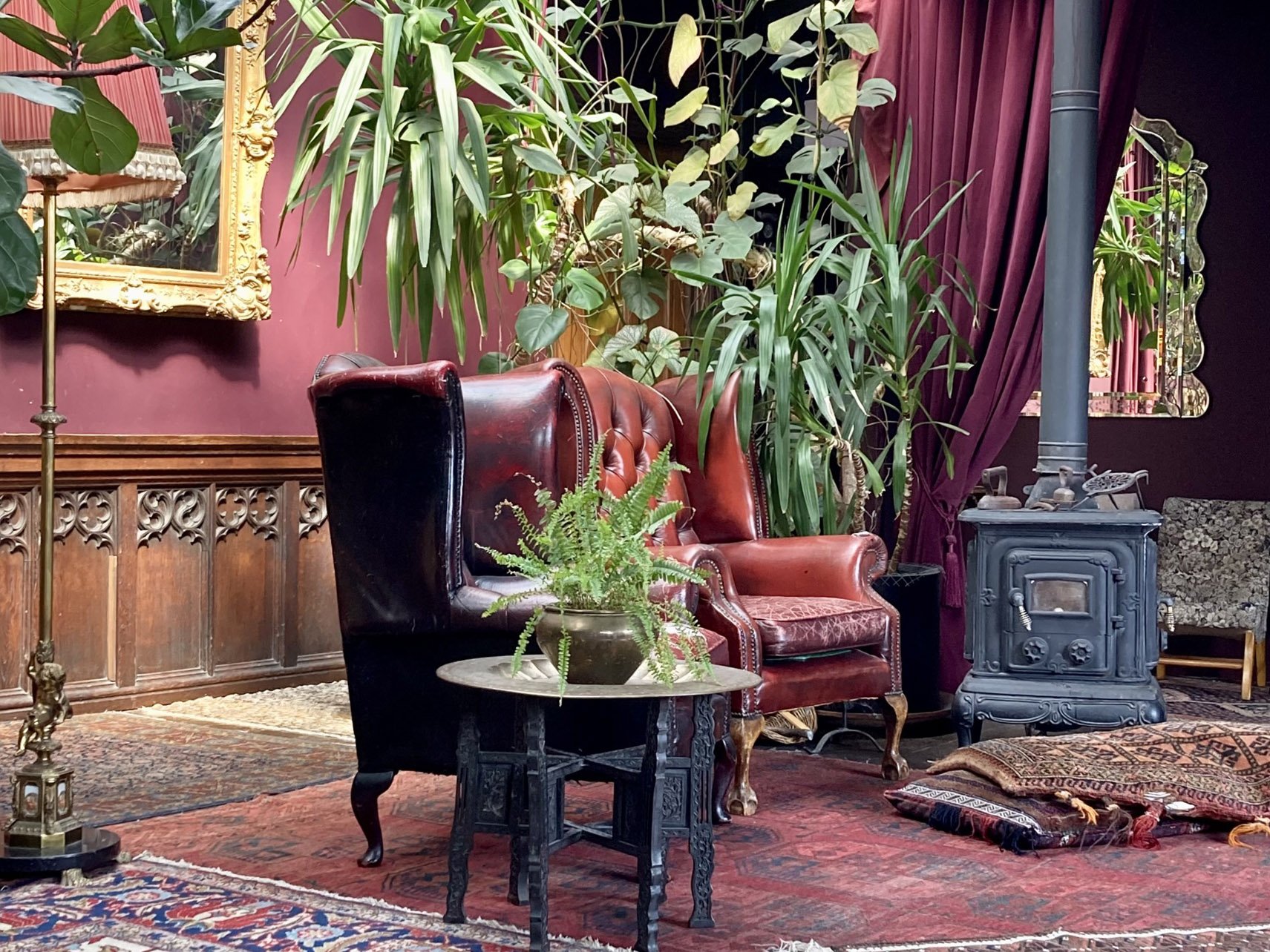 Interior of a vintage living room with a dark red wall, tall green plants, a leather armchair, a small black table with a potted fern, a wood stove, a mirror, and ornate rugs and curtains.