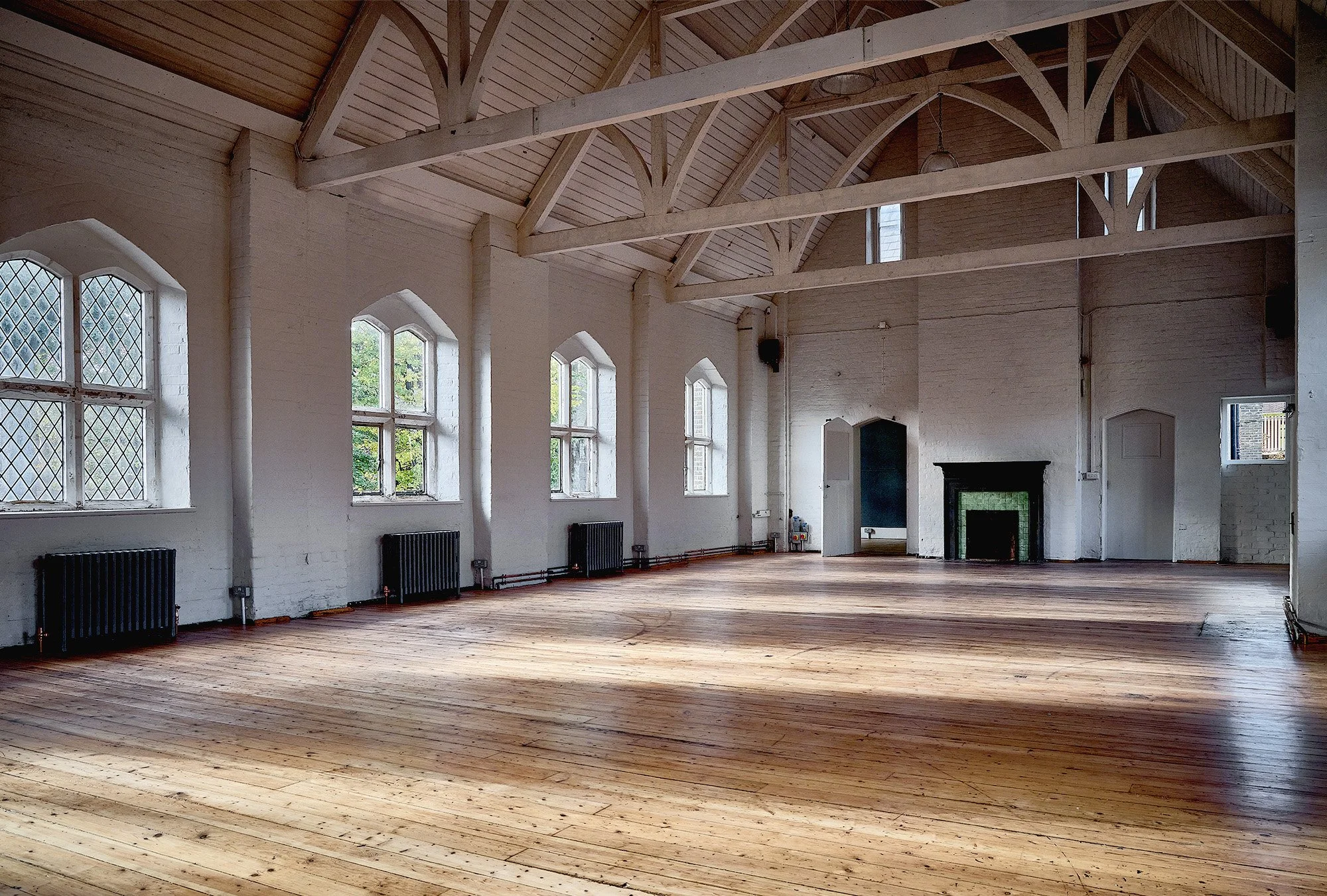 An empty spacious room with wooden floors, white brick walls, and large arched windows. The ceiling has exposed wooden beams and trusses, with a fireplace on one wall.