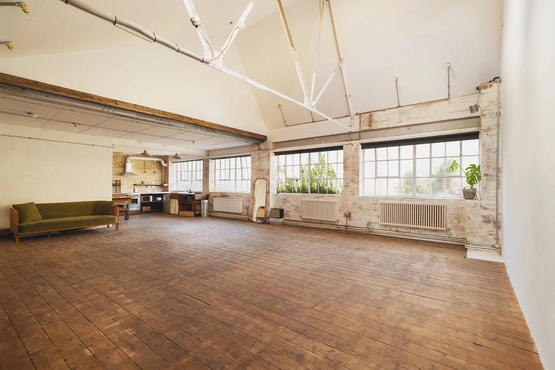 Empty loft-style room with large windows, wooden floors, a green couch, a mirror, and some plants.