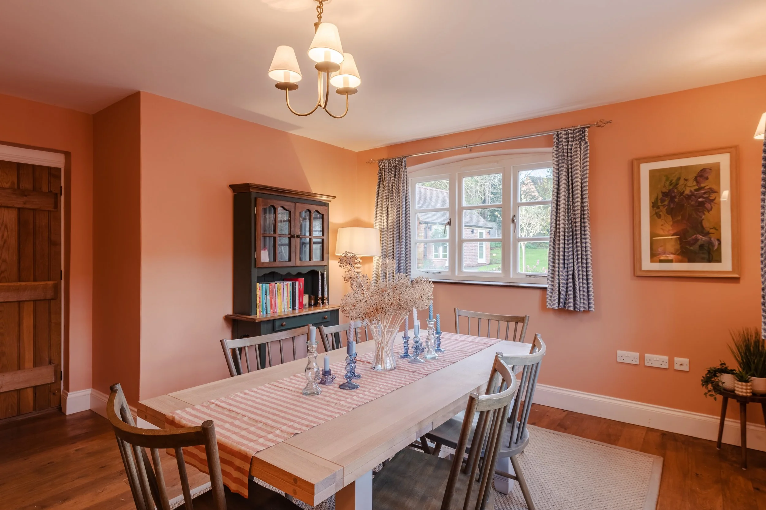 A dining room with pink walls, a wooden table with a pink and white striped table runner, candle holders, a glass vase with dried flowers, six wooden chairs, a black cabinet with glass doors and books, a window with blue patterned curtains, a framed 