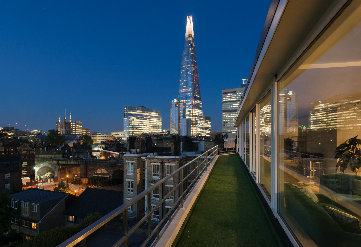 Nighttime cityscape of London with the Shard skyscraper illuminated, viewed from a rooftop terrace with a glass wall and green artificial grass carpet.