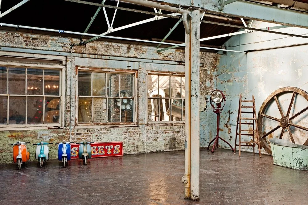 Interior of an industrial-style space with exposed brick walls, large windows, and vintage decor including a wheel, ladder, and old-fashioned lighting.