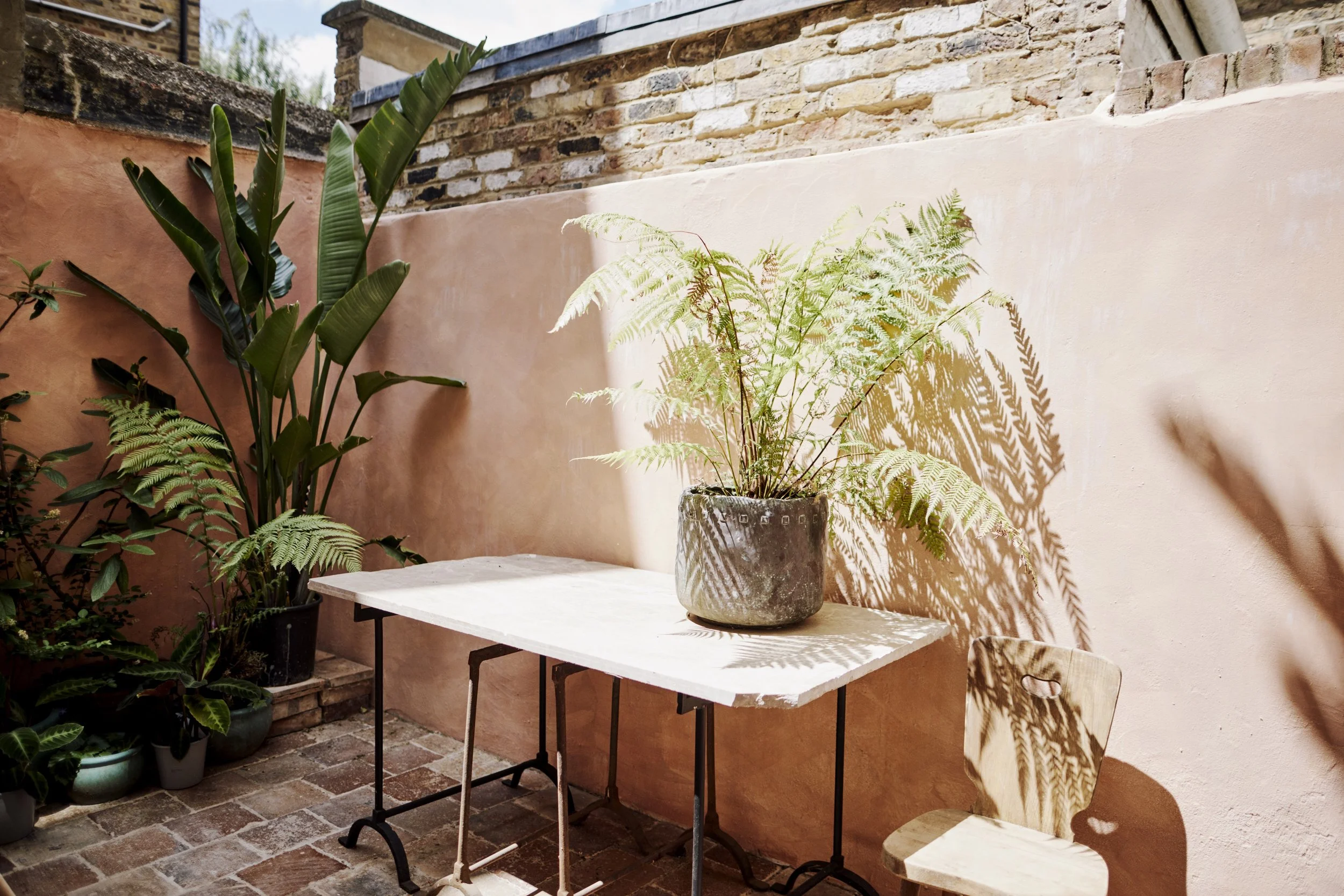 Outdoor patio with a table, a potted fern, a wooden chair, and potted plants against a pink wall, with shadows of fern leaves cast on the wall.