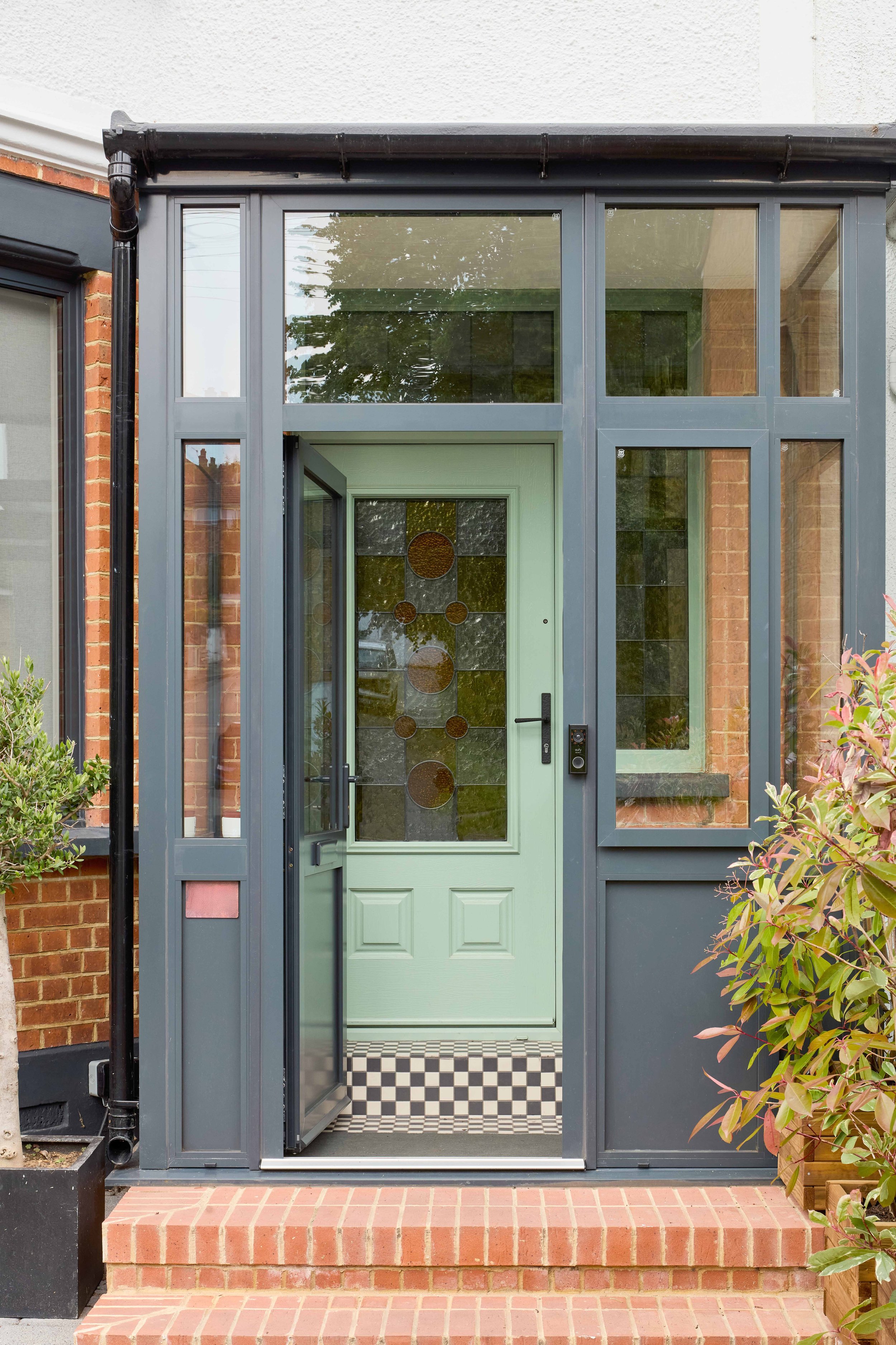 Front door with glass panels and stained glass design, surrounded by a metal porch frame, brick steps, and greenery.
