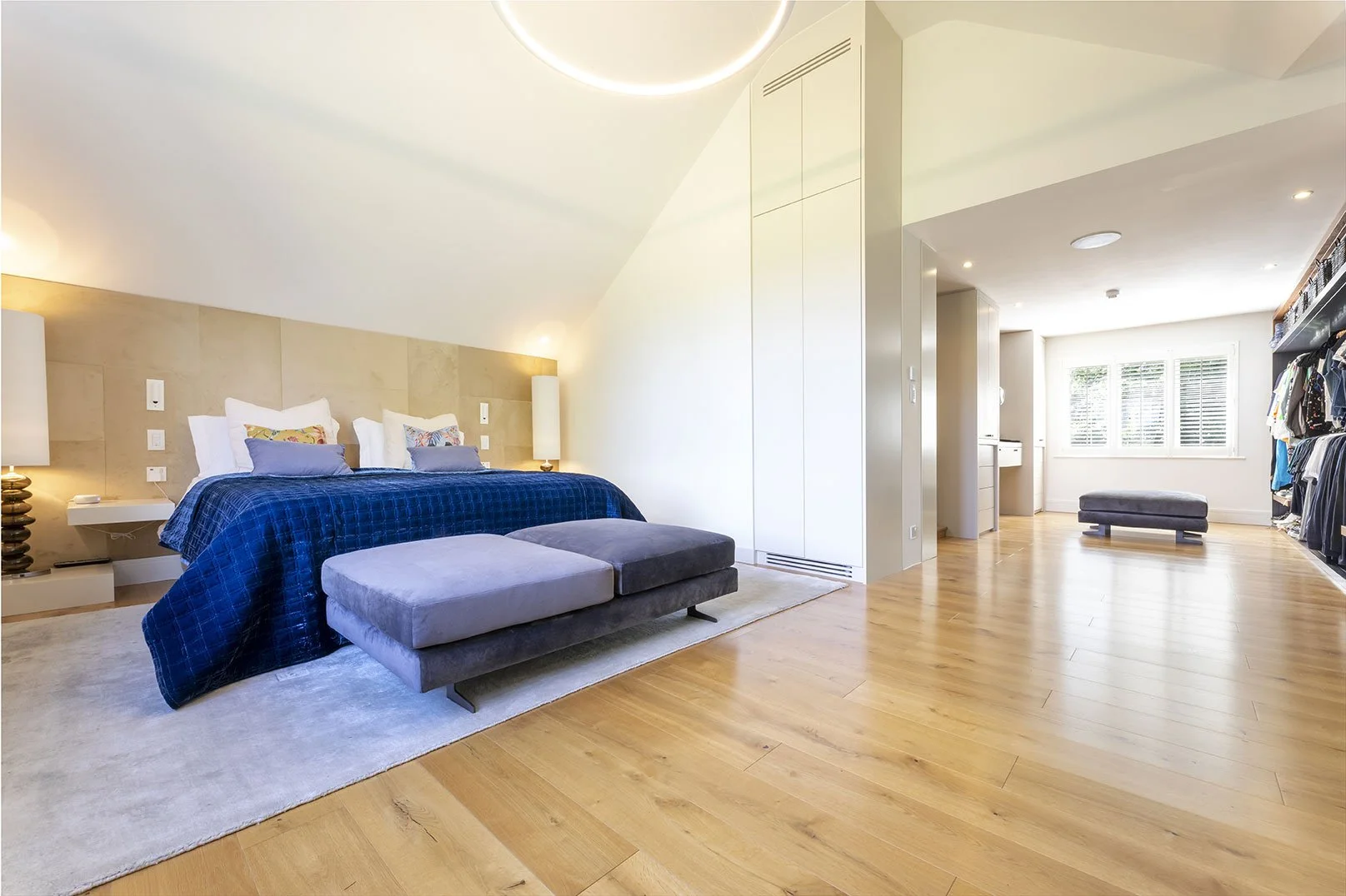 Modern bedroom with a king-sized bed, blue bedding, two bedside lamps, and a cushioned bench at the foot of the bed. A walk-in closet with hanging clothes is visible on the right, and there is a sitting area with a window in the background.