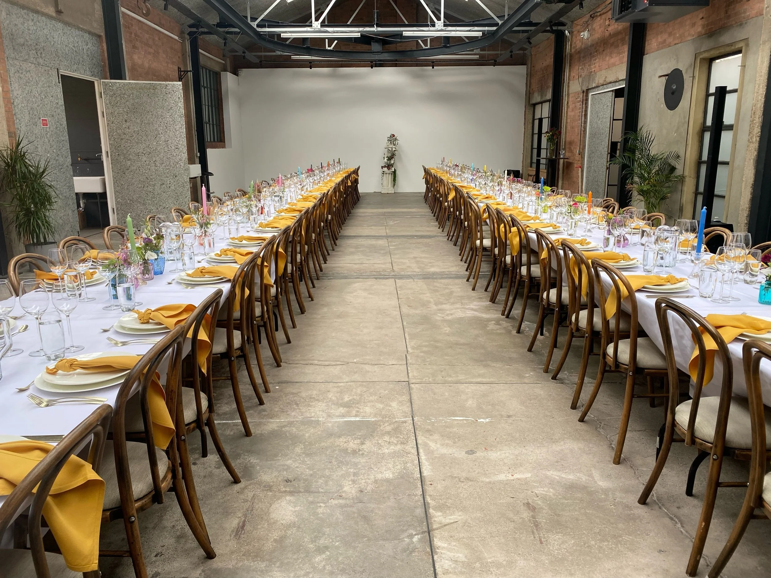 Long dining table set for a celebration or event, with yellow napkins, wine glasses, and colorful candles in a rustic industrial-style venue.