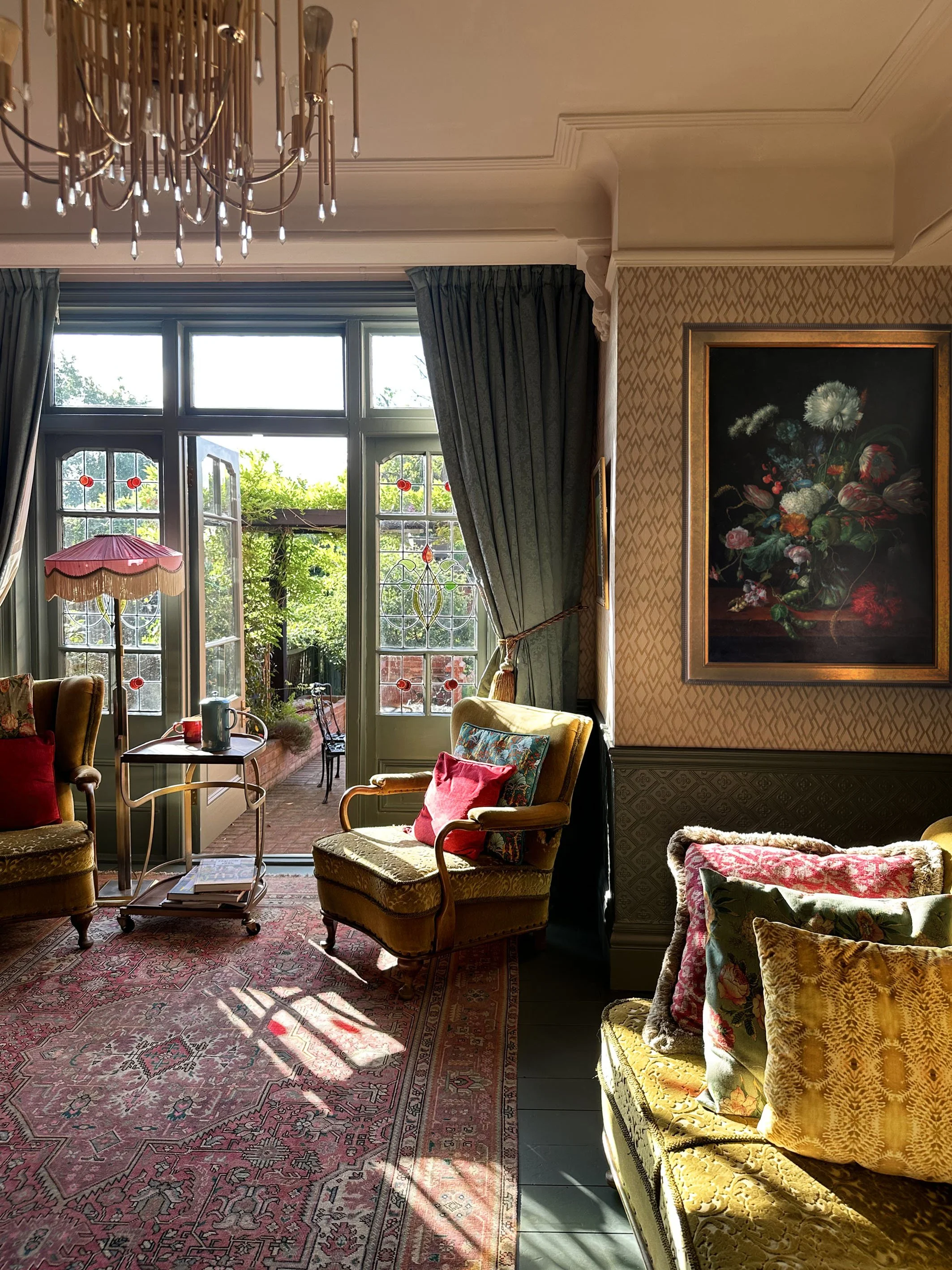 Living room with vintage furniture, colorful cushions, a patterned rug, and large windows opening to a garden. Sunlight streams inside, and decorative curtains frame the windows. A framed floral painting hangs on the wall.
