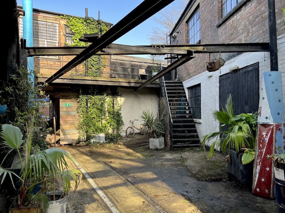 An outdoor urban scene with a concrete and asphalt ground, potted plants, a bicycle, and an outdoor staircase leading to an upper level. There is a white wall with a door, a black door or window, and a wooden structure with greenery in the background